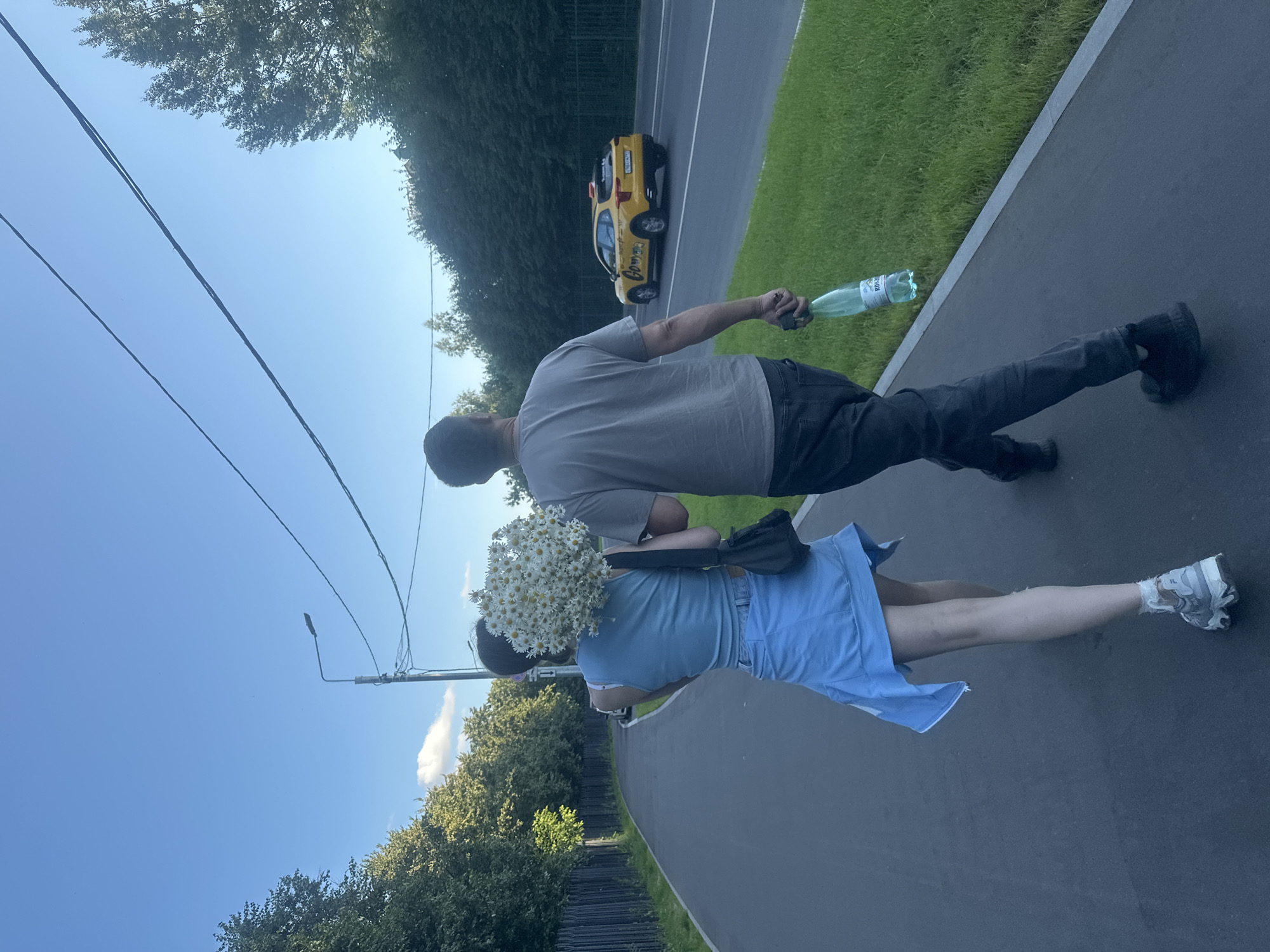 In a park-like setting in Khimki, Russia, a couple walks along a paved path. The man, wearing a grey t-shirt and dark jeans, carries a green plastic water bottle in his right hand. The woman, in a light blue dress and white athletic shoes, walks beside him, holding a large bouquet of white daisies. A black bag is slung over her shoulder. The path they are on borders a grassy area and a road with a yellow taxi visible in the distance. Trees line the background, and a lamppost stands on the left side of the image. The sky is clear and blue, suggesting a daytime setting with fair weather. The overall mood appears casual and relaxed, as the couple enjoys a stroll.