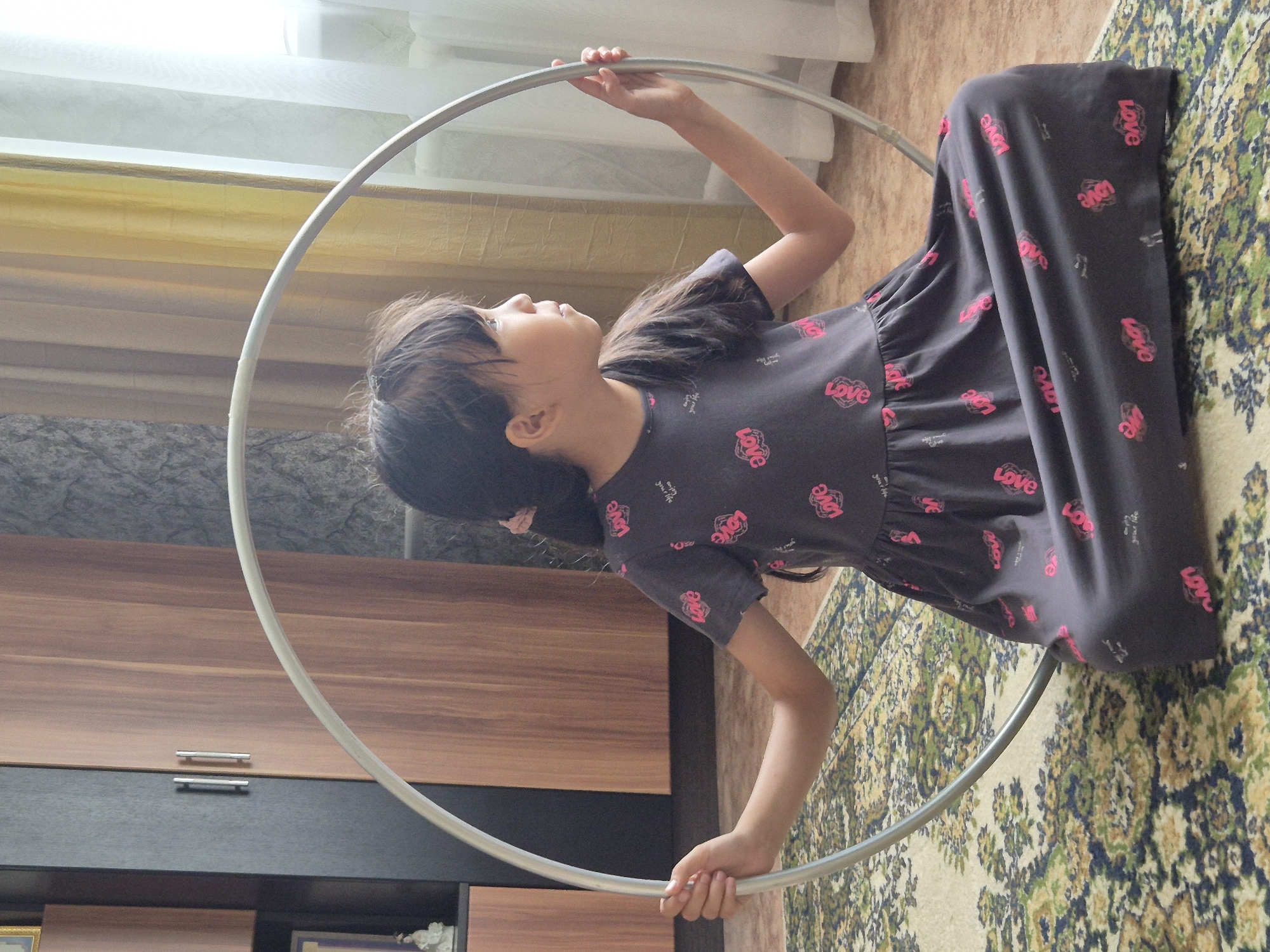 A young girl is sitting on a patterned carpet in front of a large, silver hoop. She is wearing a dark grey dress with a repeating pink "love" pattern. Her left hand rests on the hoop, and her right arm is bent. She is looking to her right, away from the camera. In the background, there is a wooden wardrobe with silver handles and sheer, yellow curtains partially covering a window. The floor is tiled. The scene appears to be an indoor domestic setting, possibly a living room or bedroom.