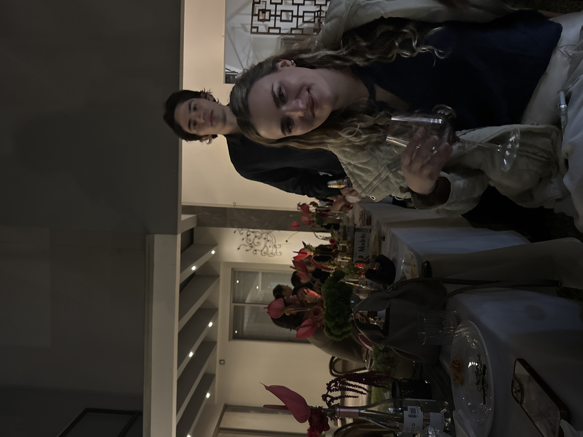 The image depicts an indoor/outdoor evening social gathering in the city of Puebla, Mexico. The scene is dominated by a long table draped with a white tablecloth, set under a ceiling featuring exposed beams and recessed lighting.
In the foreground, a Caucasian woman with long, wavy blonde hair is seated, facing the camera with a slight smile. She wears a dark top and a light-colored jacket draped over her shoulders, and holds a wine glass containing red liquid in her right hand. A silver corkscrew rests near her right thigh. Behind her, a Caucasian man with dark hair stands, looking directly at the camera. He is wearing a dark, long-sleeved collared shirt.
The table extends into the background, populated by several other individuals whose faces are largely obscured or out of focus. Numerous floral centerpieces, predominantly featuring pink anthuriums and dark red flowers, are arranged along the table in various vases and bottles. Visible objects on the table include white plates, some with residual food fragments, clear drinking glasses, an empty clear glass bottle, a light brown handbag, and a smartphone with a clear case lying face up. A bottle with "Modelo" visible on its label is positioned further down the table.
The background shows light-colored walls, a large glass sliding door or window, and decorative wall art including a dark metal scrollwork piece and a square lattice-patterned screen. The overall lighting is dim, suggesting nighttime.