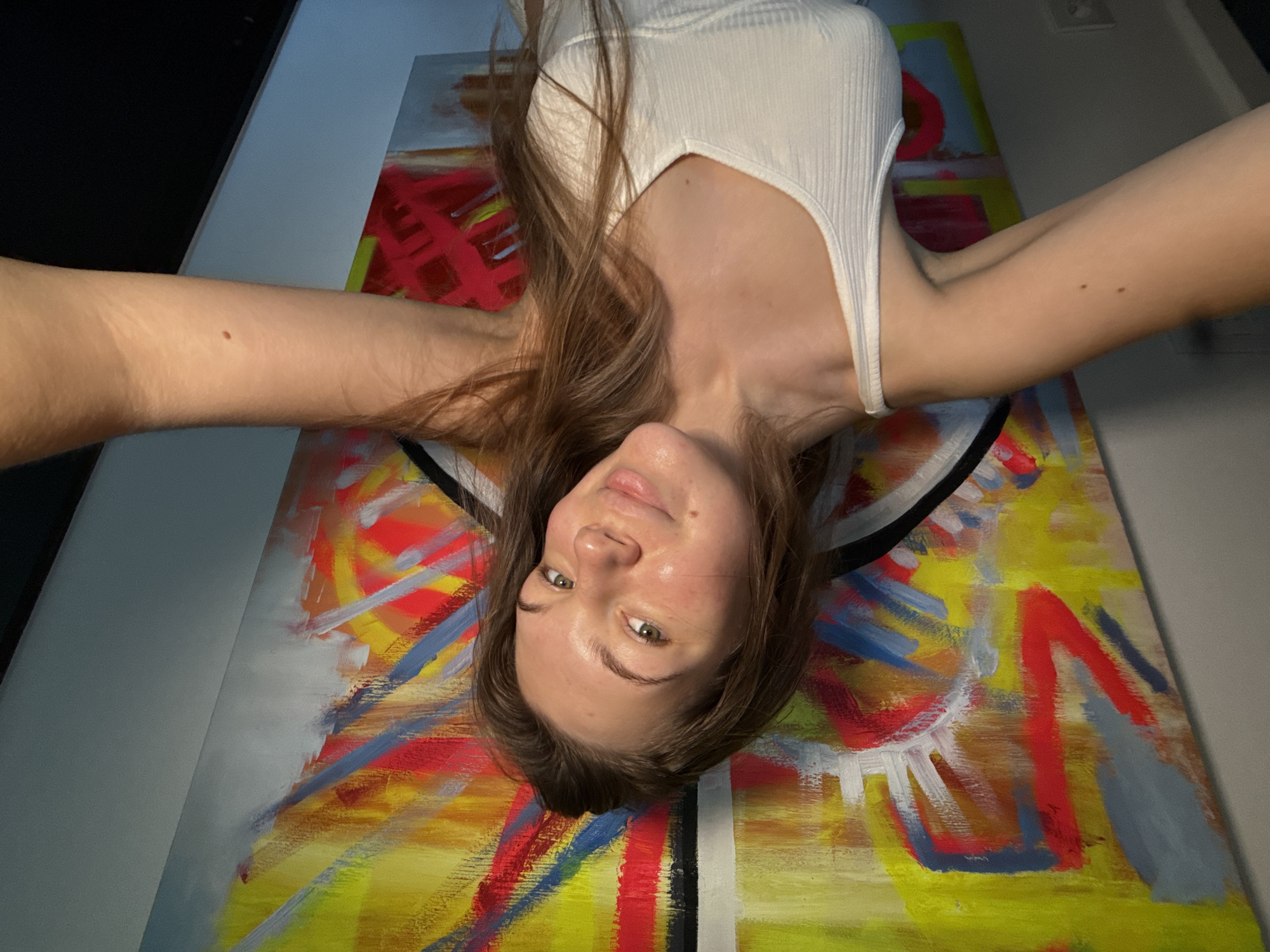 A person with fair skin and light brown hair, wearing a white tank top, is lying on an abstract painting with colors including yellow, red, blue, and white. The person is holding the camera at arm's length, taking a selfie. The background includes a gray wall and a black object in the upper left corner. The image appears to be taken indoors. The location is not discernible from the image and cannot be inferred to be Vozdovac Urban Municipality, Serbia.