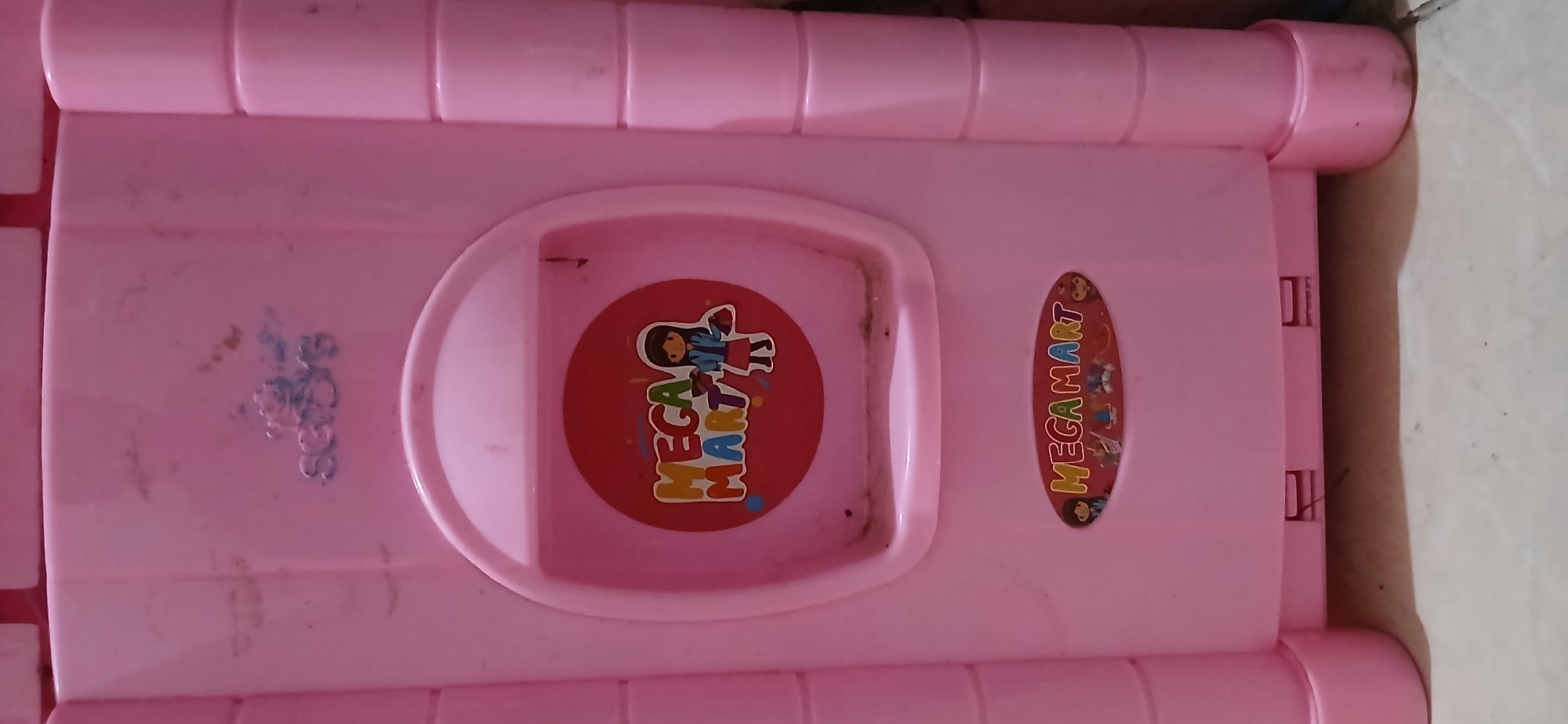 The image shows a pink plastic toy castle, likely a dollhouse or play structure, with stickers on its facade. The castle is placed on a tiled floor. A blue logo with the word "SGID'S" and a crown is visible at the top center of the castle. Below this, a circular sticker with the words "MEGA MART" in colorful letters and an illustration of a girl holding shopping bags is present. Further down, another oval sticker also reads "MEGA MART" with a collection of small illustrations including figures, shopping bags, and what appear to be toy buildings. The castle has visible dust and smudges, indicating it has been used. The overall environment suggests a domestic setting, possibly a child's room. The image does not contain information that would allow for the identification of its specific location within Samarinda, Indonesia, other than the assumption that it is a place where such toys are used or sold.