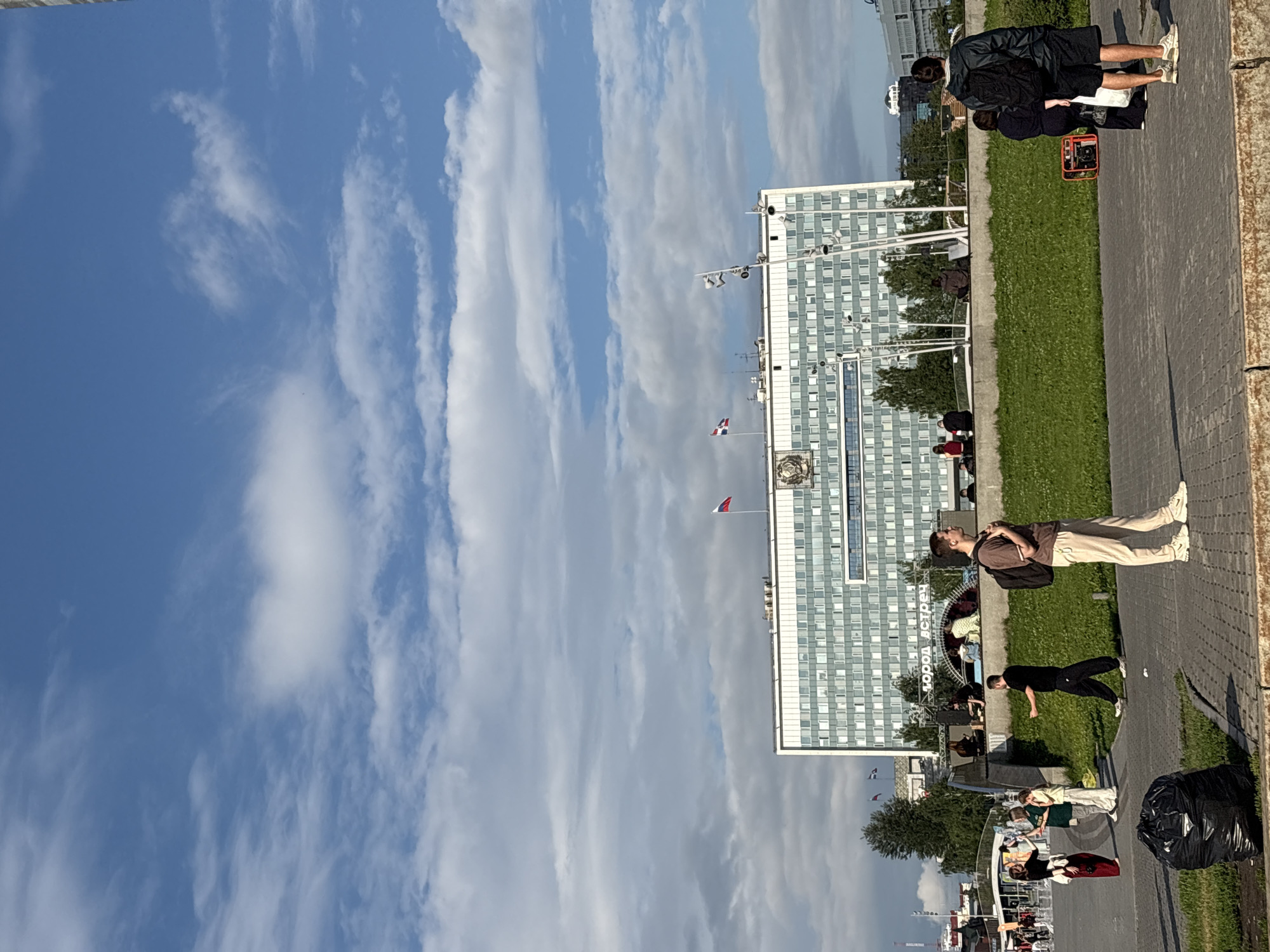 The image captures a lively urban scene in Perm, Russia, on a day with a bright blue sky dotted with fluffy white clouds. In the background, a large, modern, light-colored building with many windows dominates the skyline, identified by signage as "ГОРОД ВСТРЕЧ" (City of Meetings). Russian flags fly from the building's rooftop. In front of the building, a grassy area slopes down towards a paved plaza.
Several groups of people are present. On the grassy slope, individuals are sitting and interacting, some appearing to be watching something happening on a stage or platform. In the foreground, on the paved area, a young man with a backpack stands looking to his left. To his left, a group of people are engaged in conversation, with one woman in a red skirt and white top. Further left, a black trash bag is placed on the grass. On the far right of the frame, a person with a backpack stands, and a portable generator is visible on the grass behind them. The overall atmosphere is one of a public gathering or event, with people enjoying the outdoors.
