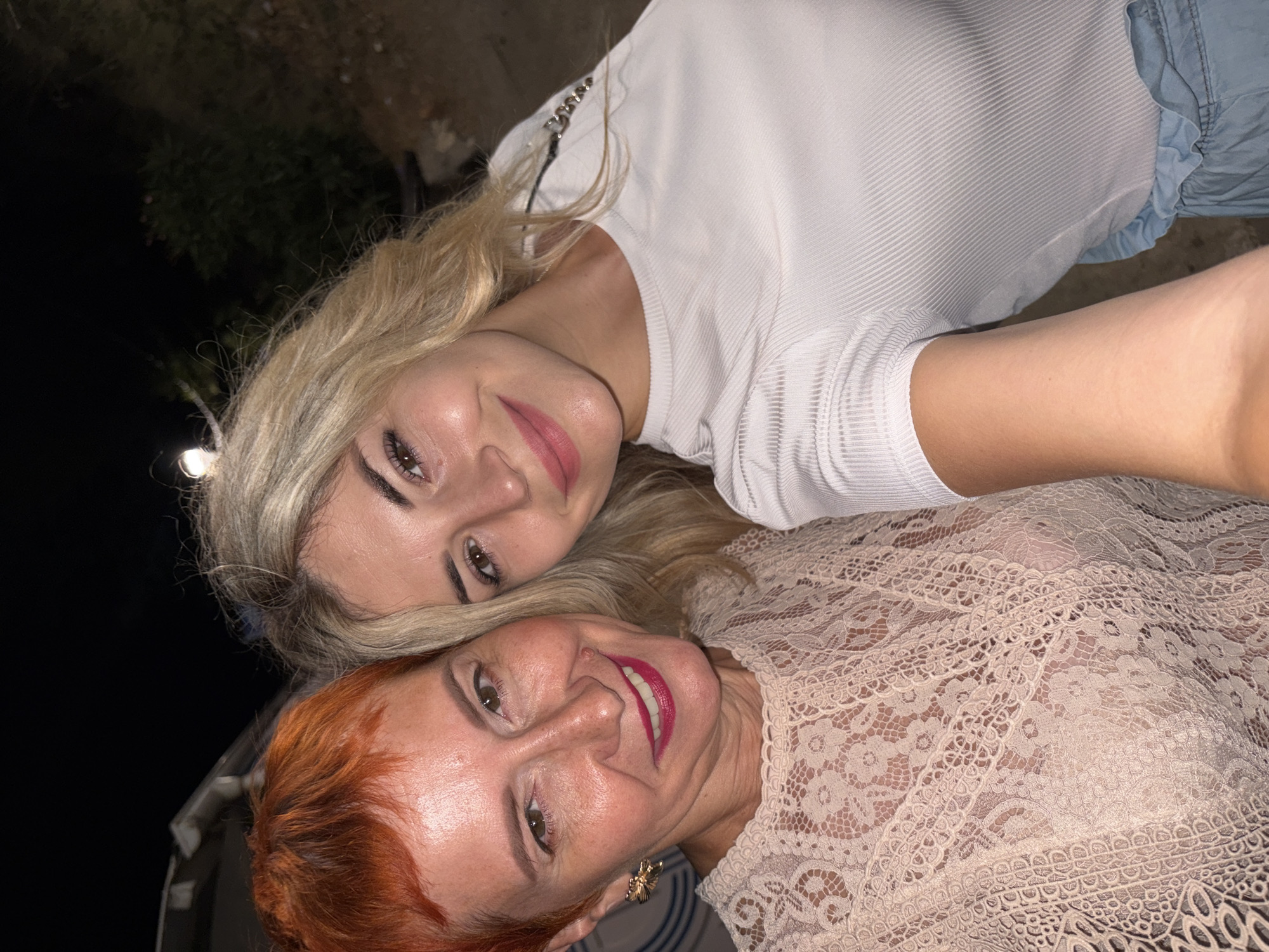 The image depicts a close-up selfie of two women. The woman on the left has short, vibrant orange hair and is wearing a light-colored, intricately patterned lace top and a gold bee-shaped earring. She has bright pink lipstick on and is smiling. The woman on the right has long, blonde hair and is wearing a white ribbed t-shirt. She is also smiling, with her head tilted slightly. The background is dark and appears to be outdoors at night, with some greenery and structures visible. The overall setting suggests a casual social gathering. While the image is identified as being in Budva, Montenegro, no specific city landmarks or context are discernible from the visual information alone.