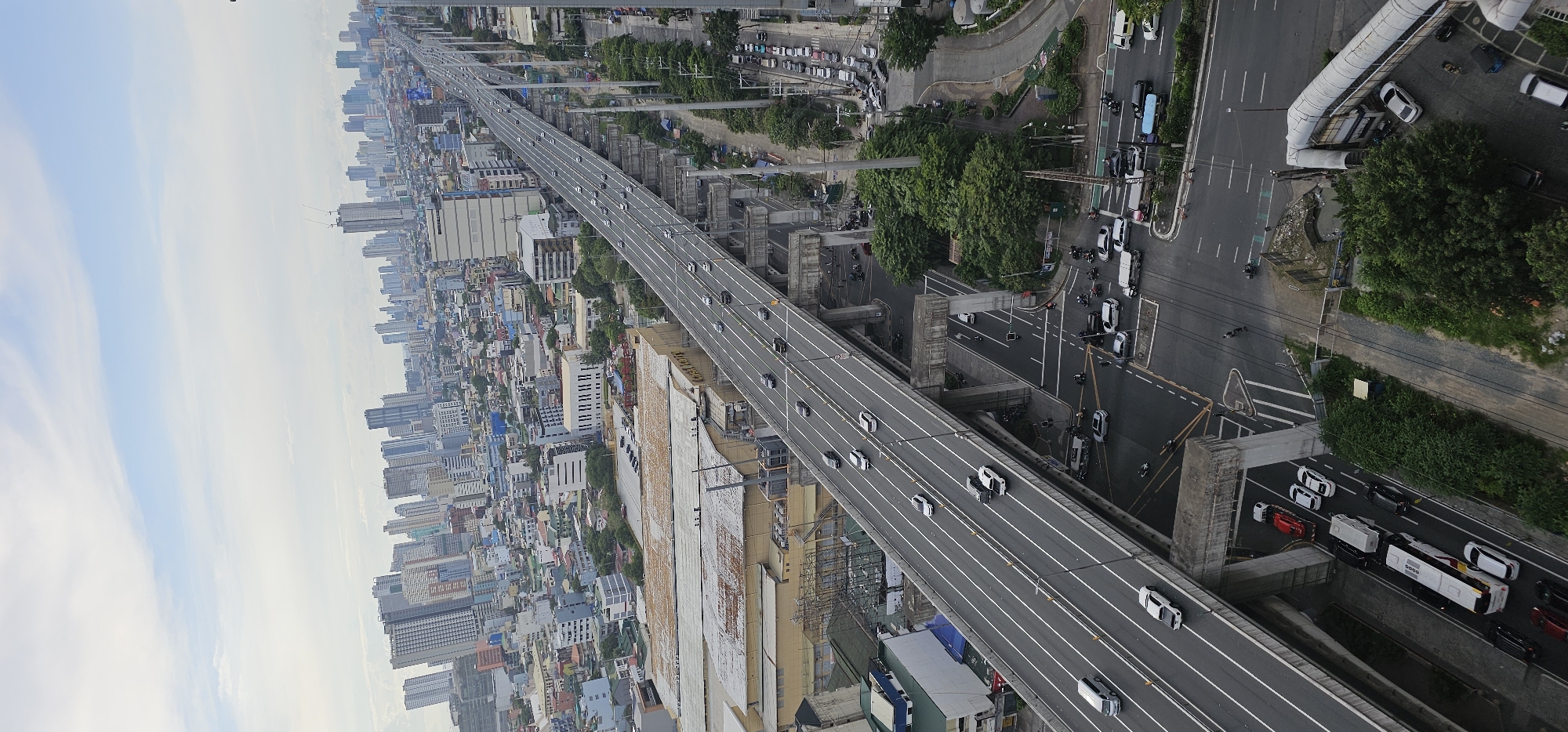 This is a high-angle, vertical shot of a multi-lane elevated highway passing through a dense urban environment in Makati City, Philippines. The sky is overcast with scattered clouds. The highway is populated with a moderate flow of vehicles, including cars, vans, and trucks. Below the highway, various buildings are visible, including residential structures, commercial buildings, and what appears to be a large warehouse or industrial facility with a corrugated metal roof. Trees and vegetation are interspersed throughout the landscape. At street level, beneath an overpass, a traffic intersection shows several vehicles and motorcycles, indicating a busy roadway. The overall impression is of a modern, bustling metropolis with extensive infrastructure designed to manage significant traffic volume.