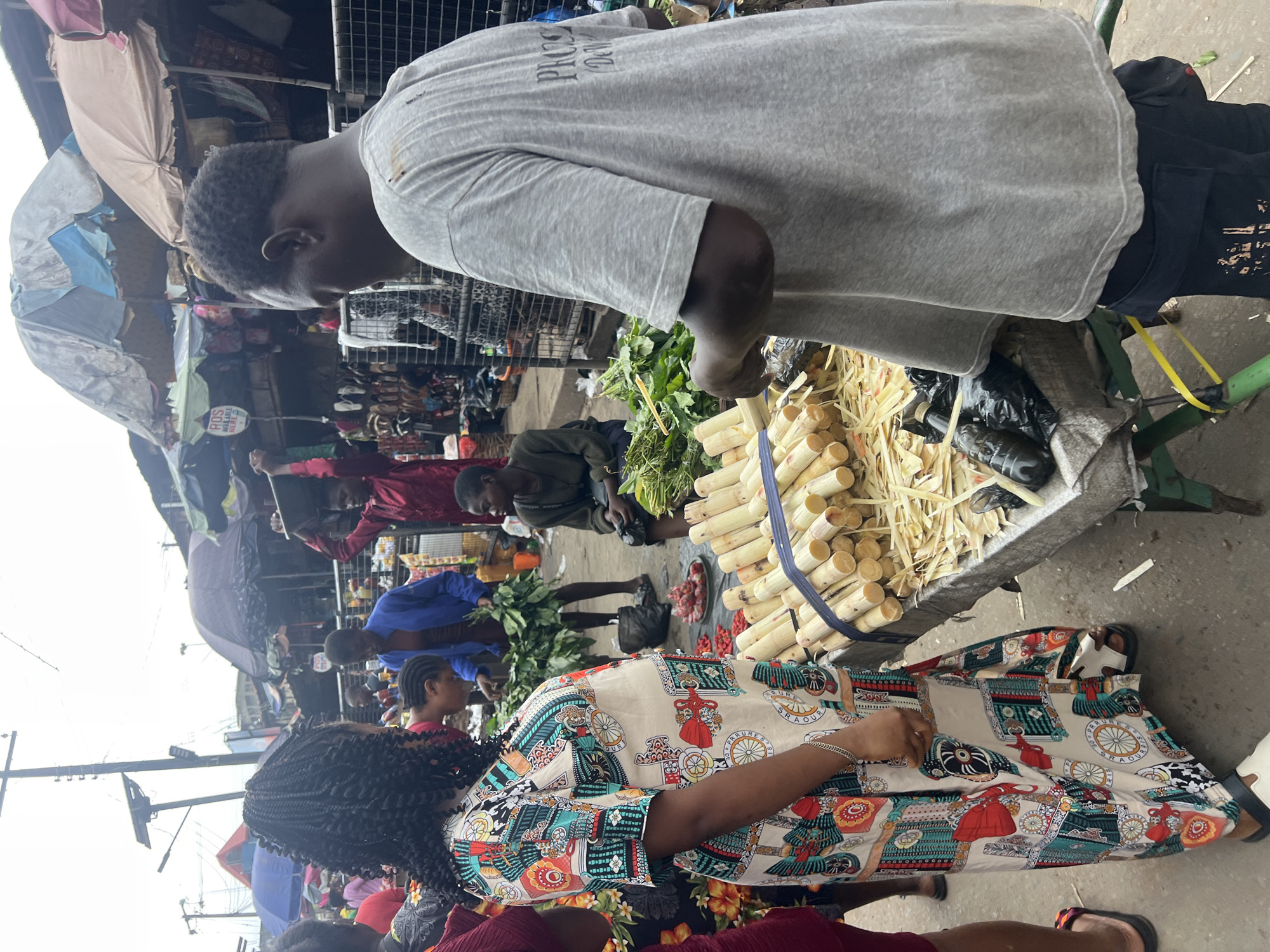 In Nkpor, Nigeria, a bustling open-air market scene unfolds. The foreground is dominated by a woman, viewed from the side, wearing a vibrant, multi-patterned dress. She stands near a cart laden with stalks of sugarcane, some neatly bundled with blue strapping and others already cut and piled on a table. Beside the sugarcane, a man in a grey t-shirt and dark pants is engaged in what appears to be food preparation or transaction, with black plastic bags and more produce visible. Behind them, other market-goers mill about. To the left, a person in a bright red top carries a large basket on their head. Other individuals are seen interacting or tending to their stalls, under the shelter of various tarpaulins and umbrellas. The ground is paved and appears somewhat wet in places. The overall atmosphere suggests a busy commercial hub within the city.