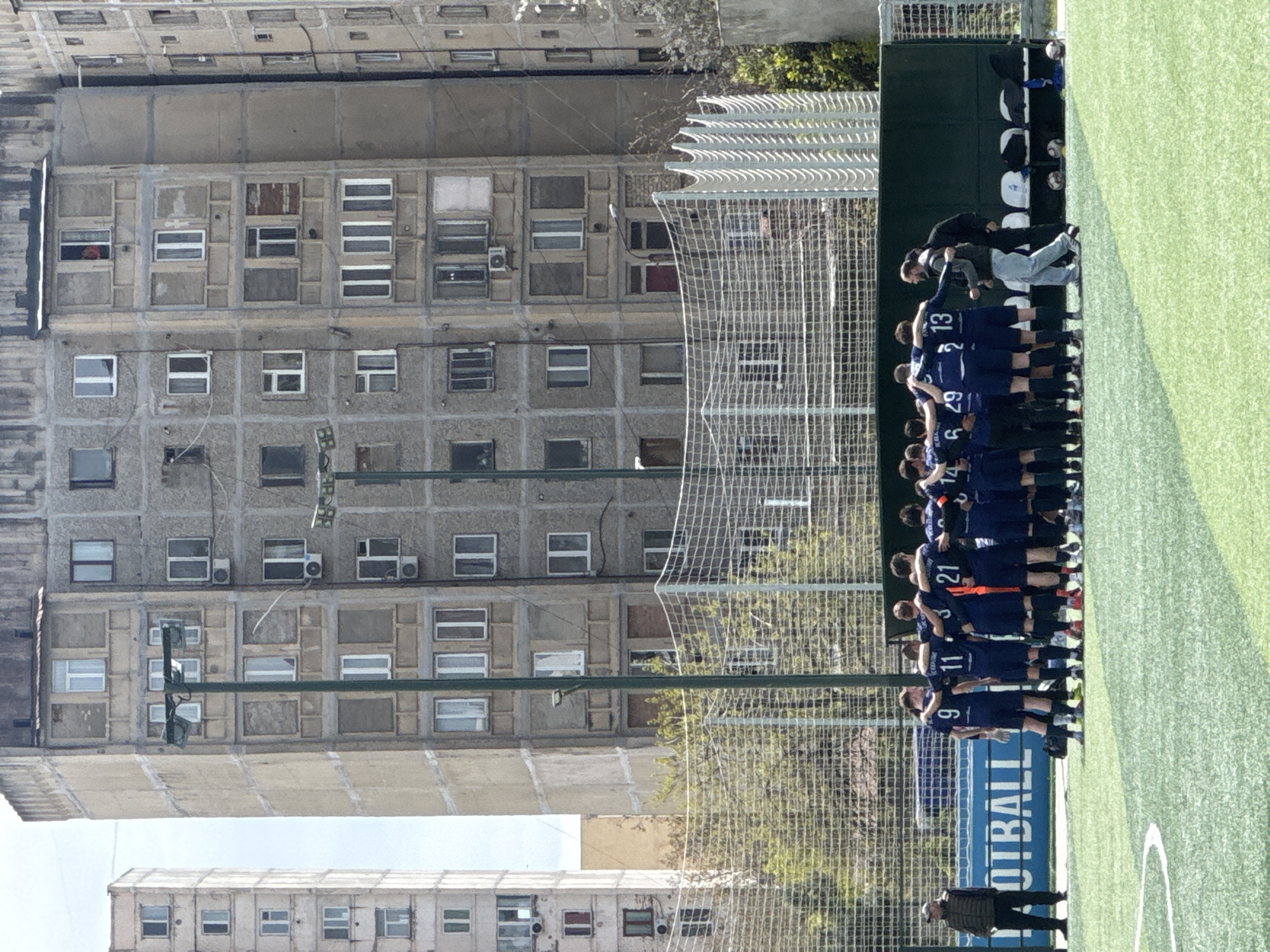 A wide shot of a soccer match on a green field adjacent to a large, grey, concrete apartment building with multiple windows, some equipped with air conditioning units, in Tbilisi, Georgia. A soccer team wearing navy blue uniforms and white numbers is standing together on the field. Behind them is a green wall with the word "FOOTBALL" in large white letters. A man in a dark jacket and hat is walking along the edge of the field. A net fence separates the field from the building.