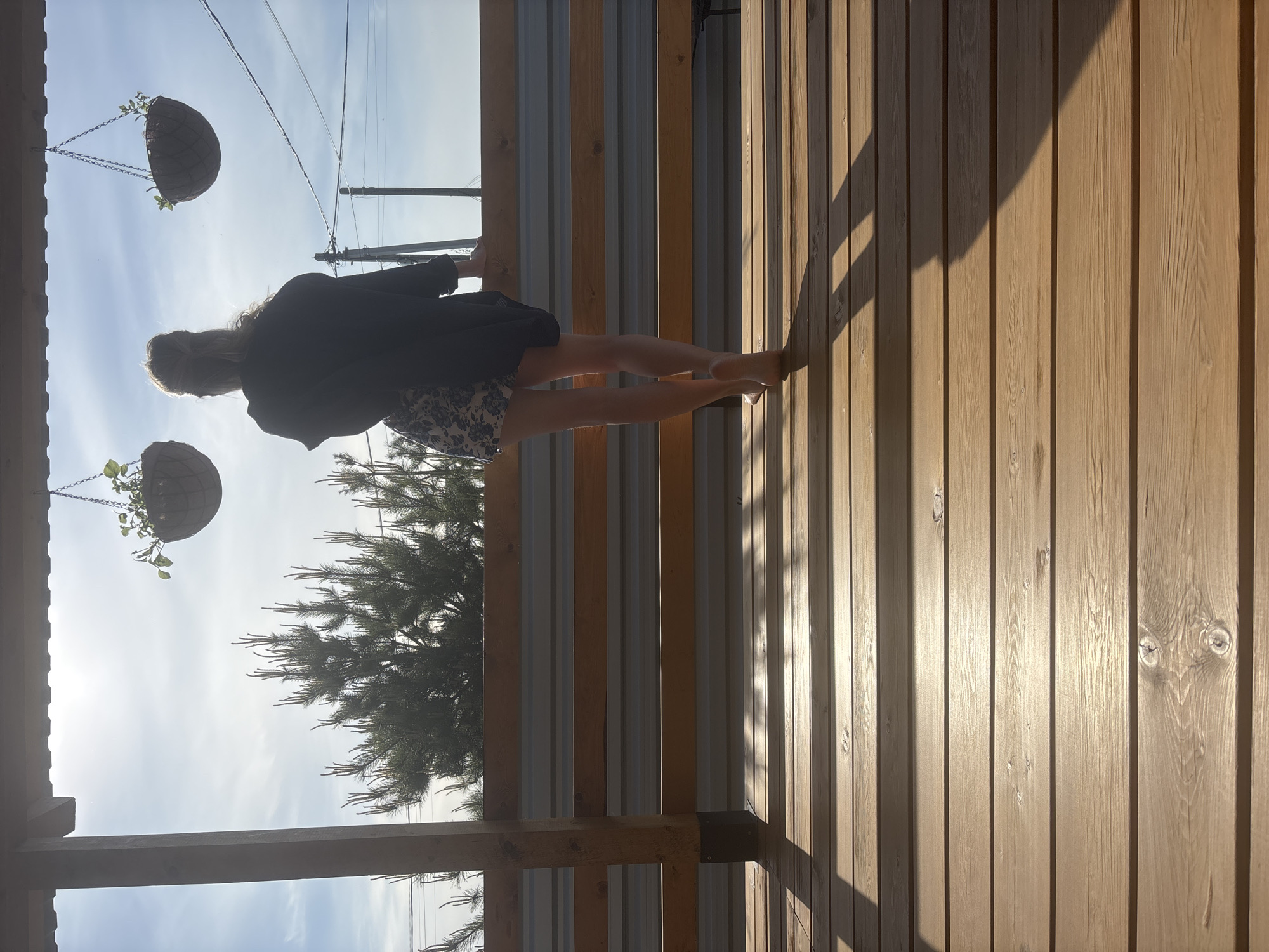 The image depicts a woman standing on a wooden porch in Chaikovsky, Russia. She is wearing a dark blazer over a floral dress. The porch has a wooden floor and a metal railing with wooden slats. Hanging planters are visible overhead. A pine tree is visible in the background. The sky is visible in the background, with power lines stretching across it. The scene is bathed in sunlight.