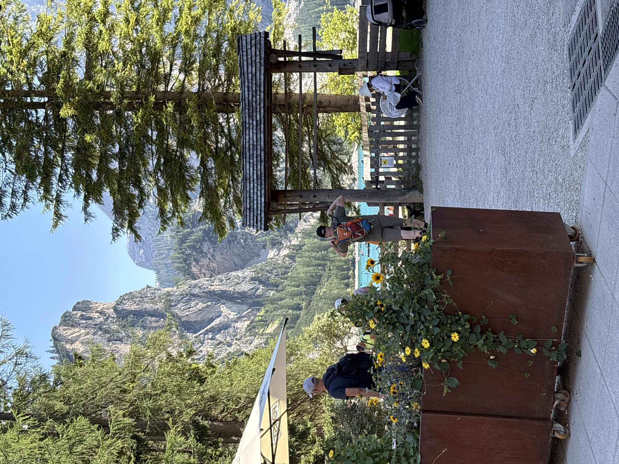 The image depicts a scenic view of Lake Braies in Prags - Braies, Italy. In the foreground, a large, rustic, rust-colored planter box is filled with vibrant yellow flowers and green foliage. To the left, a man wearing a gray cap and a backpack stands under a large, cream-colored umbrella. Behind him, partially obscured by greenery, another person is visible.
In the middle ground, a wooden archway with a shingled roof stands prominently. Beyond the archway, a rustic wooden fence encloses an area where a woman in a white hat sits on a chair, reading a document. To her right, a stroller is parked. A man wearing a cap and a colorful backpack is positioned near the archway, holding up his phone as if taking a photograph.
The background reveals the breathtaking scenery of Lake Braies, with its turquoise waters and surrounding steep, forested mountains. The sky is clear and blue, indicating a sunny day. The overall scene captures a serene and picturesque moment at a popular tourist destination.