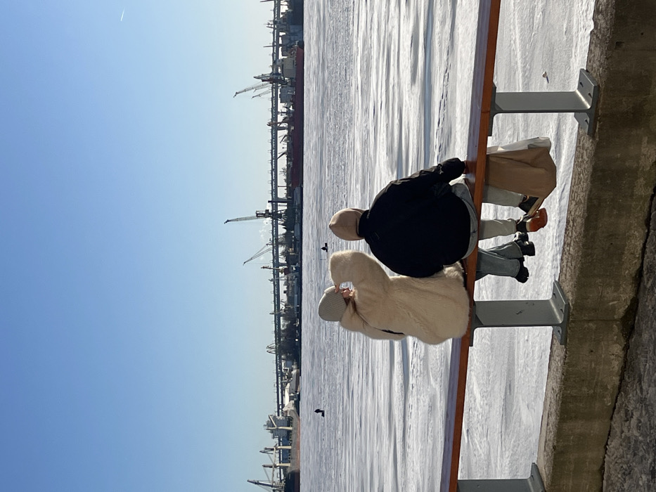 Two individuals, observed from behind, are seated side-by-side on a wooden railing on an embankment overlooking a wide, frozen body of water under a clear blue sky. The scene is located in Saint Petersburg, Russia, likely along the Neva River or Gulf of Finland, facing an industrial port area.
The individual on the left wears a light-colored, possibly cream, textured coat and a light beanie, with blue jeans and dark shoes. Their left arm is raised, hand near their forehead, suggesting they are shielding their eyes or looking intently into the distance. The individual on the right wears a dark, possibly black, hooded jacket with the hood up, light-colored pants, and dark shoes with visible reddish-brown soles. A light brown paper bag hangs from their left side.
The frozen water, covered in white ice and snow, extends into the midground. Several distant figures are visible on the ice, appearing to be ice fishing. The background features a large industrial port with multiple cargo ships, numerous tall cranes (including gantry and mobile cranes), and other port infrastructure. A long, multi-span bridge, likely part of the Western High-Speed Diameter, spans the water behind the port facilities. The overall environment suggests a cold, clear winter day.