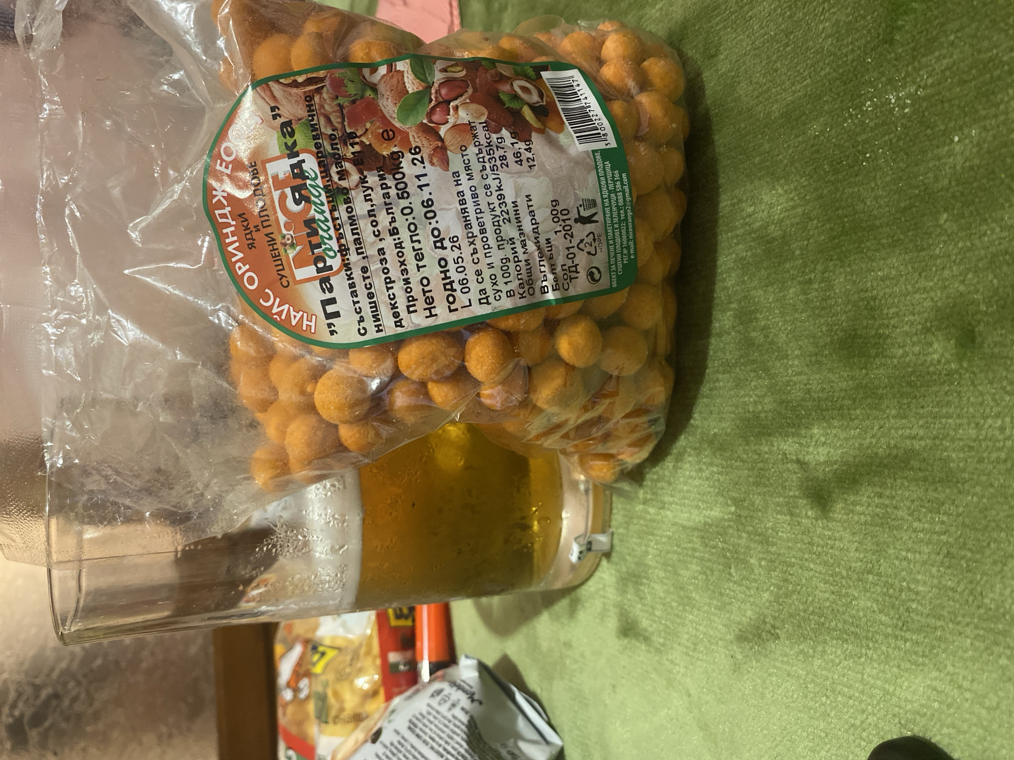 The image displays a close-up of a glass of beer next to a plastic bag of orange-coated peanuts. The bag has a label with Bulgarian text that reads "Най-с ориндж бо", which translates to "Most Orange Bo". The label also includes nutritional information and a date of "06.11.26". In the background, another snack package is visible. The setting appears to be indoors, with a green, textured surface in the foreground. The image does not provide any specific information about the city of Smolyan, Bulgaria, or any people.
