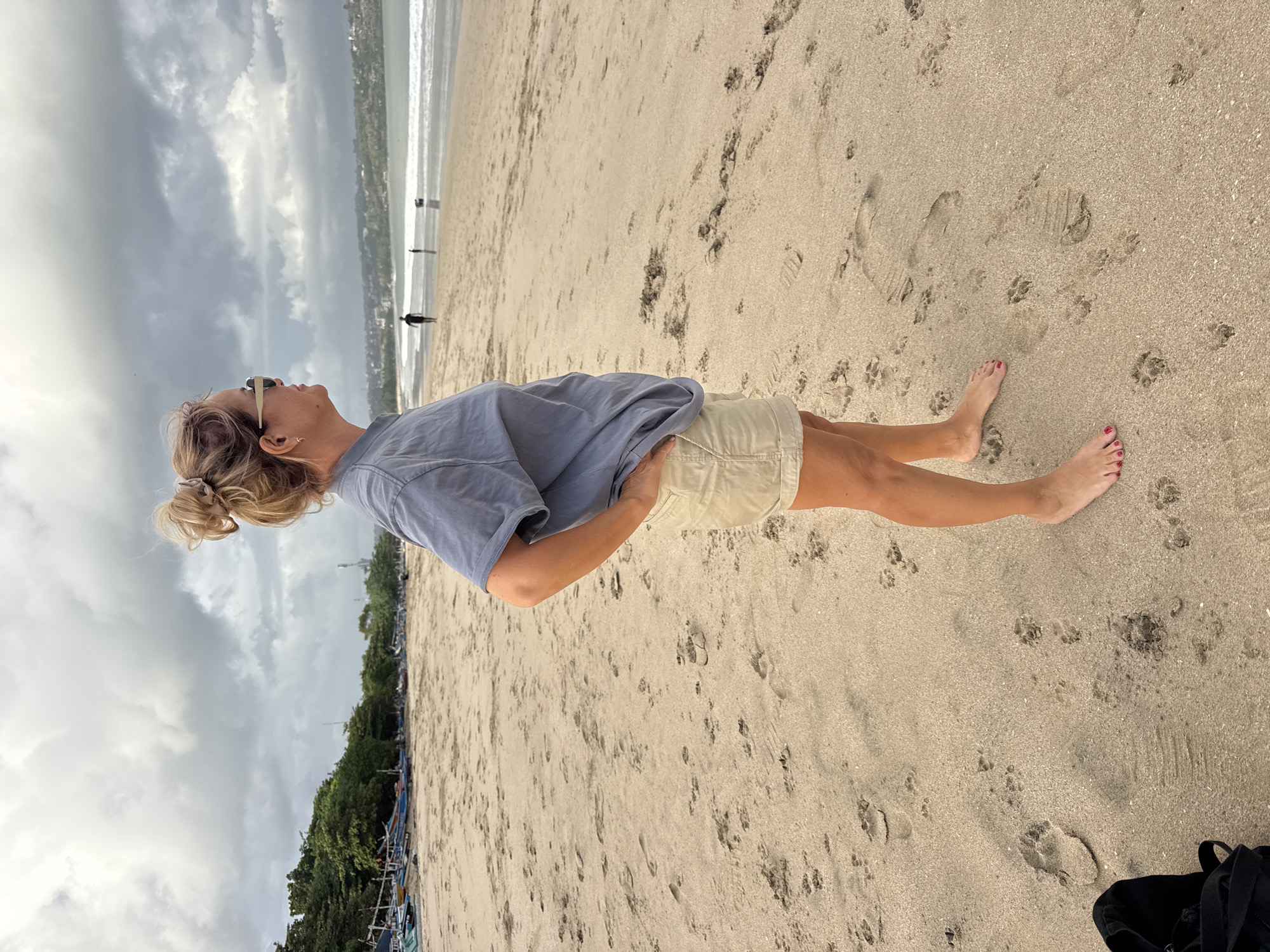 A woman stands on the sandy beach in Kuta, Indonesia, facing the ocean. She has blonde hair pulled into a bun and wears sunglasses, a blue t-shirt, and beige shorts. Her hands are placed on her hips. The beach is mostly empty, with a few footprints in the sand and some boats lined up in the background. The sky is overcast, with dark clouds suggesting impending rain. In the distance, a few people can be seen walking along the shoreline and a hotel can be seen in the background.