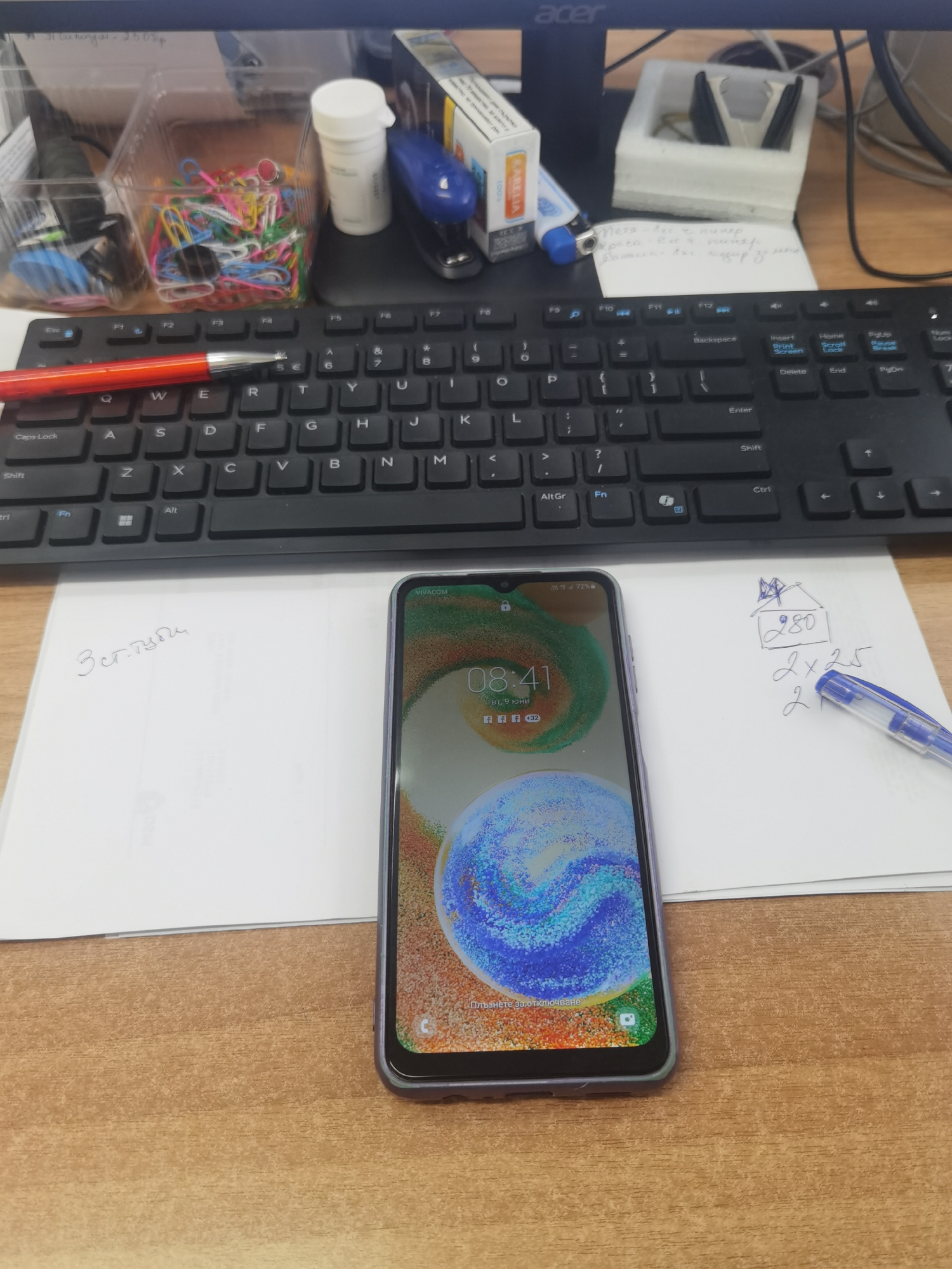 The image depicts a close-up, eye-level view of a wooden desk surface. In the foreground, a smartphone with a purple case rests on a white piece of paper. The phone screen displays the time as 08:41 and indicates the date as 9th June. Next to the phone, also on the paper, is a handwritten note with a drawing of a house and some mathematical calculations: "280", "2 x 25", and "2". A blue pen lies on the paper, next to the handwritten calculations. Behind the paper, a black computer keyboard is visible, with a red pen resting on it. Further back, there are office supplies including a stapler, a bottle of white pills, and a clear plastic container filled with colorful paper clips. A computer monitor with an "acer" logo is at the top of the frame. The scene appears to be an indoor office or workspace. There is no indication of the city of Lomets, Bulgaria, in the image.
