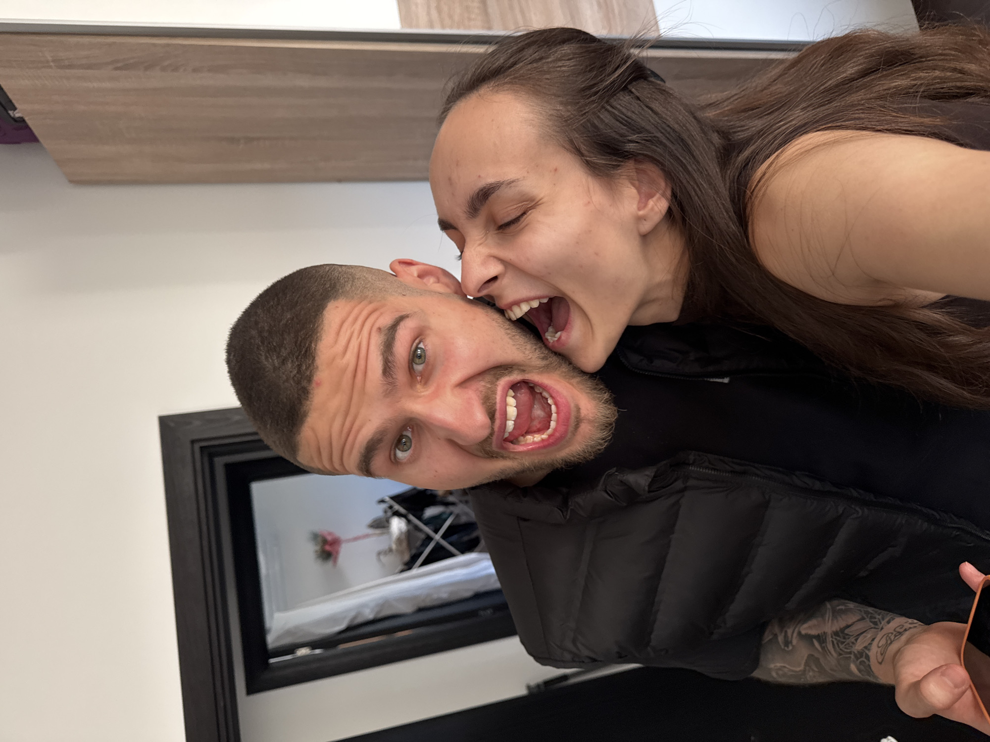 The image captures a rotated close-up of two individuals, a man and a woman, engaged in a playful interaction. The woman, positioned on the left side of the frame, has long dark hair and is wearing a dark top. Her face shows an exaggerated expression, with her eyes squinted or nearly closed, and her mouth wide open, appearing to playfully "bite" or nip the man's ear/cheek area. Her teeth are visible.
The man, on the right side of the frame, has short dark hair, a beard, and is wearing a black puffer vest over a dark shirt. He is reacting with an equally exaggerated expression, his mouth wide open in a theatrical scream or laugh, revealing his tongue and teeth. His eyes are wide open, looking towards the camera. Tattoos, featuring intricate designs, are visible on his left arm, extending below the short sleeve of his vest. He is holding a phone with a distinctive orange or pink case, partially visible in the bottom right corner of the image.
The background is minimally in focus. At the top, a wooden-patterned surface, possibly a shelf or cabinet, spans horizontally. Below the man's head, a black-framed mirror or picture frame reflects an indoor setting, showing what appears to be a bed or mattress with white bedding, and some other indistinct objects. The surrounding walls are plain white.
The scene depicts a moment of lighthearted fun and humor between the two subjects, captured indoors. There is no visible text in the image. The location context indicates it was taken in Sofia, Bulgaria.