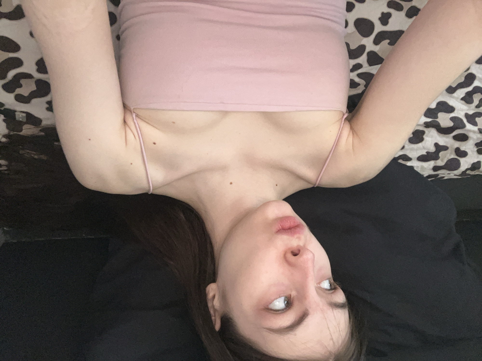A woman is lying on her back in what appears to be an indoor setting. She is wearing a light pink tank top with thin straps. Her arms are raised above her head. She has long brown hair, fair skin, and is looking to the side with her lips puckered. The background behind her consists of a patterned fabric or bedding, possibly with an animal print (such as leopard or cow print). Below her, there is a solid black fabric. The location cannot be confirmed as Saint Petersburg, Russia, based on the visual information provided in the image.
