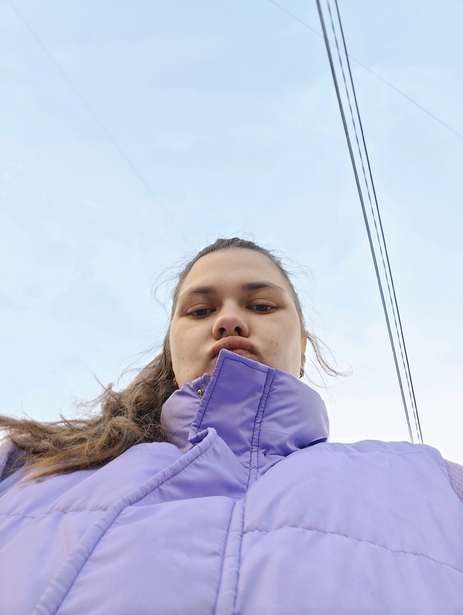 A low-angle close-up shot depicts a woman with brown hair wearing a light purple quilted vest, standing outdoors. Overhead power lines are visible against a pale blue sky. The woman's face is partially obscured by the angle, but her eyes, nose, and pursed lips are visible. The setting appears to be an urban area. Specific geographic location of Chaikovsky, Russia, is not discernible from the image.
