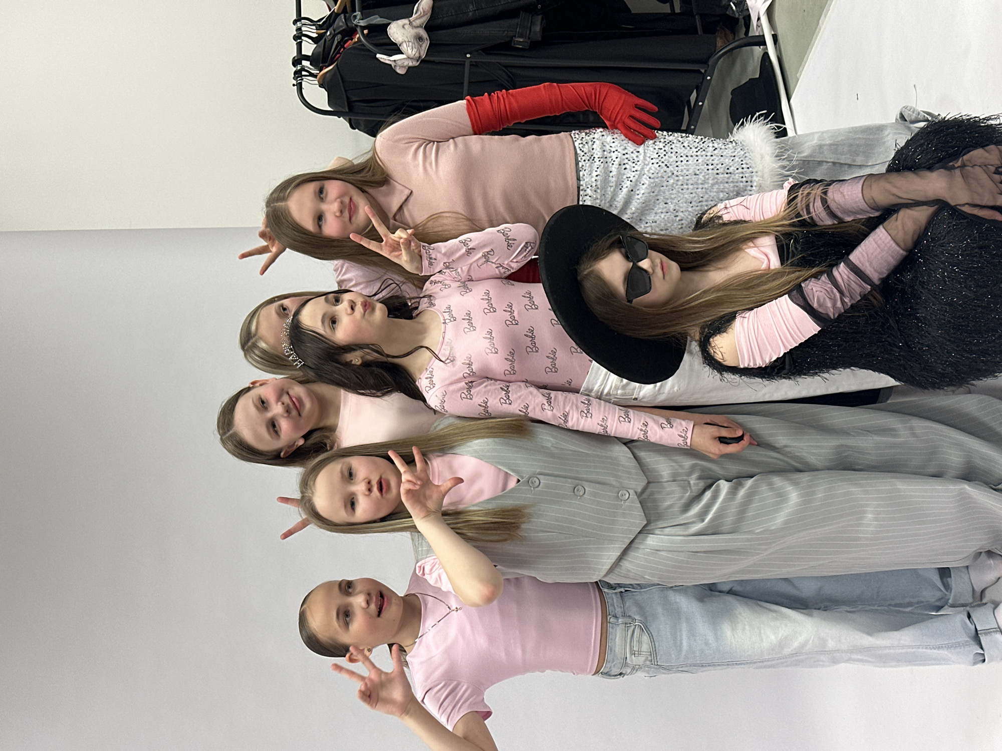 A group of six young girls are posed together in what appears to be a studio or backstage area, possibly in Votkinsk, Russia, given the prompt context. They are stacked vertically, each lying down in a row. The girls are wearing different outfits in shades of pink and gray, with some incorporating elements of "Barbie" style, such as clothing with "Barbie" printed on it. They are making peace signs with their fingers and smiling. The girl at the top is wearing red gloves and a sequined skirt with white trim. One girl is wearing a wide-brimmed black hat and sunglasses. In the background, there is a rack of clothing. The environment is a plain studio space, with a white backdrop.