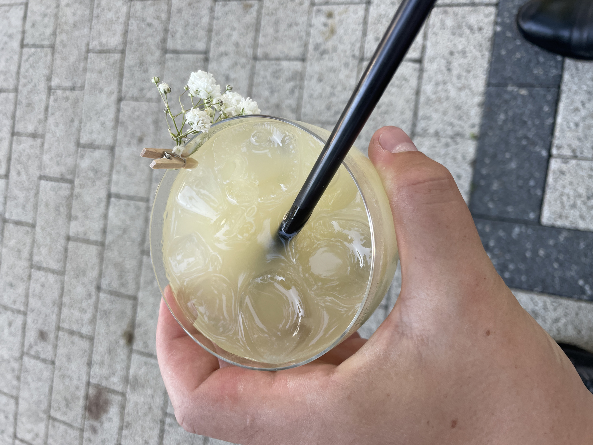 In Ratuș, Moldova, a person's hand holds a clear glass filled with a pale yellow, ice-filled beverage. A black straw protrudes from the drink, and a small, wooden clothespin secures a sprig of tiny white flowers to the rim of the glass. The scene is set against a backdrop of grey brick paving stones, indicative of an outdoor urban setting.