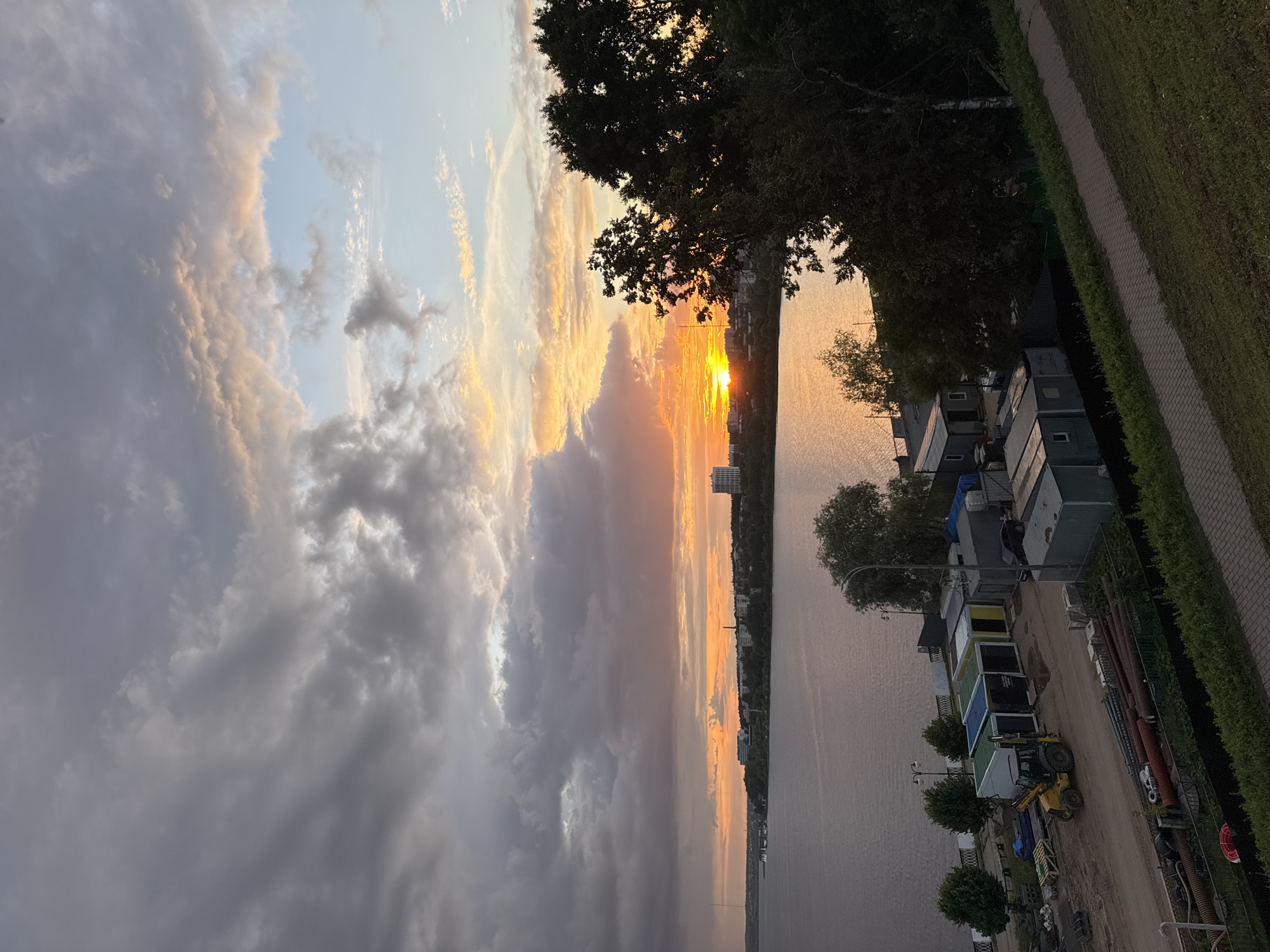 This is a vertical, candid, full-shot photograph of a waterfront scene at sunset. The sun, a bright orange orb, is low on the horizon, casting a warm glow across the sky and water. The sky is filled with dramatic clouds, ranging from dark gray to soft pink and golden hues, indicating a dynamic weather system.
On the right side of the image, a row of portable buildings, possibly used for storage or temporary housing, lines a dirt path. A small yellow construction vehicle is parked near these buildings. Beyond the buildings, a dense line of trees creates a dark silhouette against the vibrant sky. A paved walkway with a grassy border runs along the edge of the scene. The water appears calm, reflecting the colors of the sunset. In the distance, along the far bank of the water, a cityscape with several tall buildings is visible.
The overall mood is serene and contemplative, with the natural beauty of the sunset contrasted by the presence of human-made structures. The lighting suggests it is late evening, with the sun just beginning to dip below the horizon. No text is visible in the image.