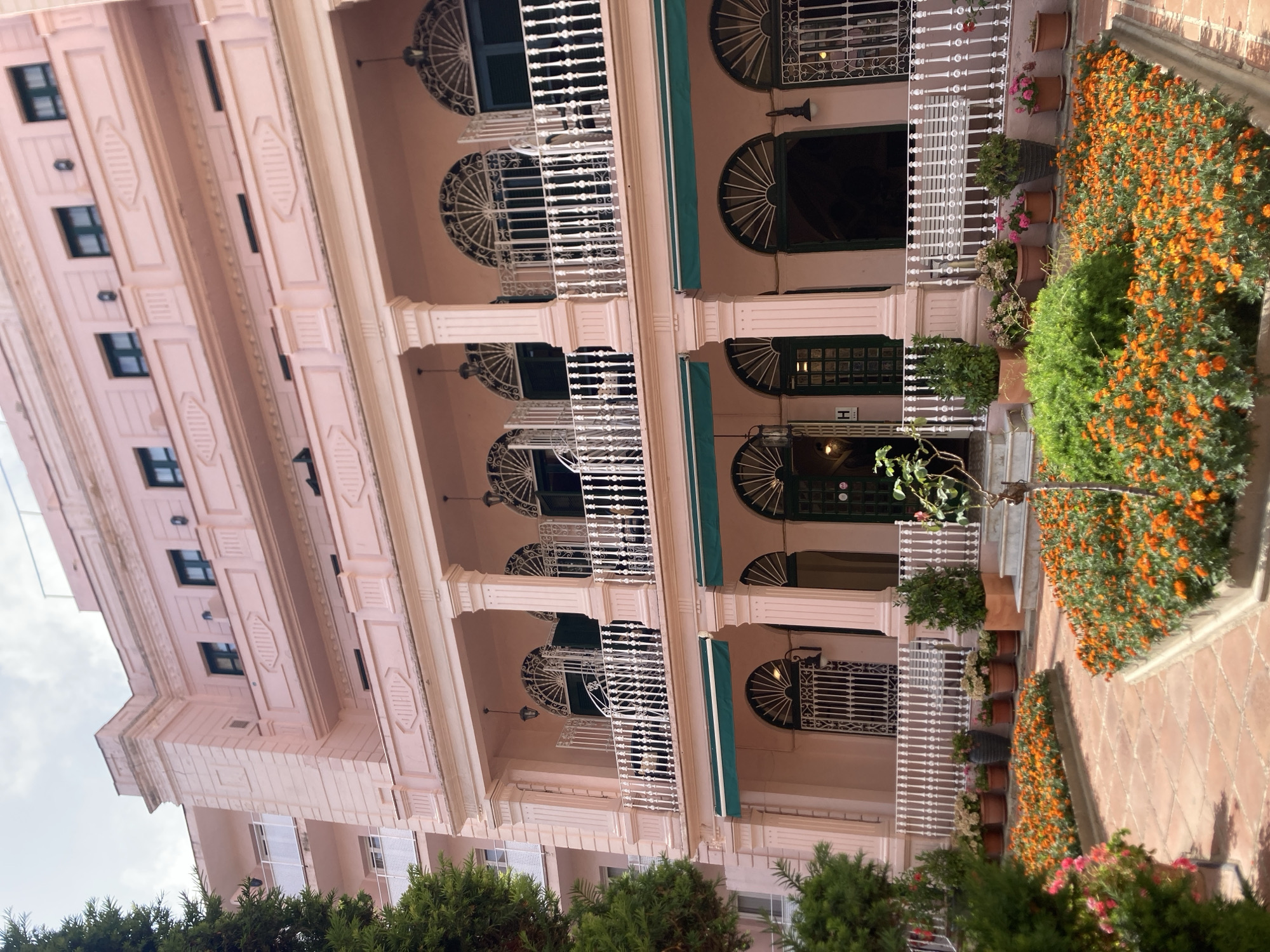 The image shows a pink building with ornate balconies and arched windows, likely a hotel or apartment building, in Lloret de Mar, Spain. A courtyard in front of the building is filled with bright orange flowers and lush green plants. Terracotta tiles pave the courtyard, and several potted plants line the balconies and courtyard. The architecture suggests a classic European style, possibly with a Mediterranean influence, consistent with the coastal town of Lloret de Mar. The sky is cloudy, indicating a potentially overcast day. There are no people visible in the scene, and the focus is on the building's facade and the vibrant courtyard garden.