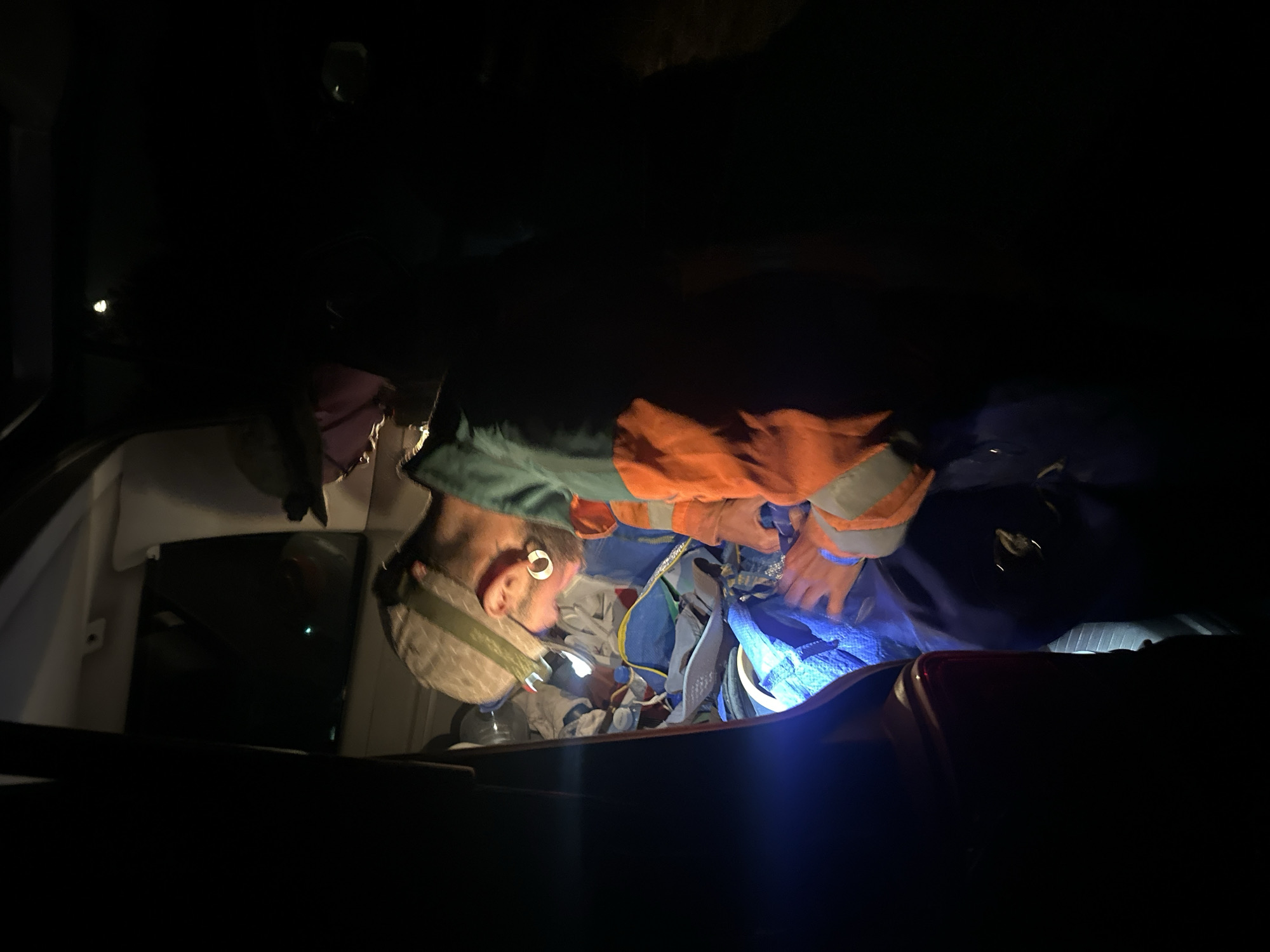 In the dimly lit interior of a vehicle, two individuals are seen interacting with supplies. One person, wearing a patterned cap, green and orange high-visibility clothing, and gloves, appears to be handling blue fabric. A bright light emanates from the left side of the image, illuminating a portion of the vehicle's interior and the contents. The scene takes place at night, with the surrounding environment not visible. The location is specified as Bijele Poljane, Montenegro.