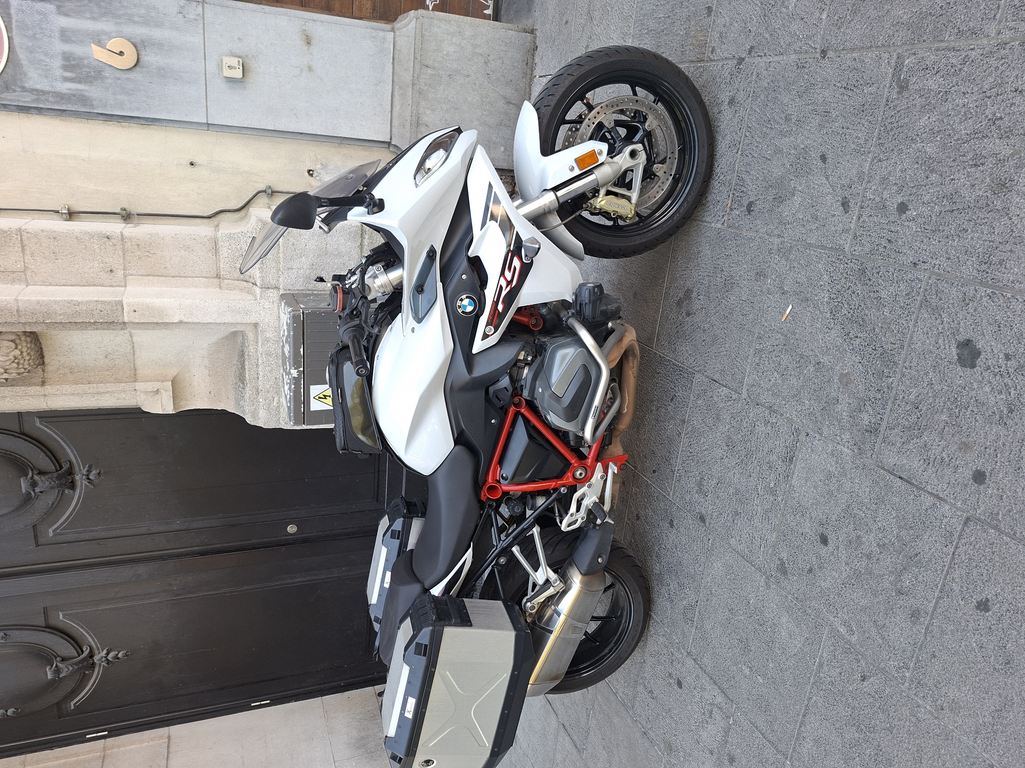 A white BMW R1250RS motorcycle with silver side cases is parked on a stone-tiled sidewalk in front of a large, dark-wood double door. The motorcycle has a red frame and a black seat. The scene appears to be in a city, likely in Europe, given the architecture and the presence of a European-style building number "6" on the wall to the right. The lighting suggests it is daytime. The background includes a portion of a stone building with decorative elements and a metal conduit running down the facade. The sidewalk is made of large, grey stone tiles with some dark spots and a discarded cigarette butt near the rear wheel of the motorcycle. The overall environment is urban and appears to be a street or a passage.