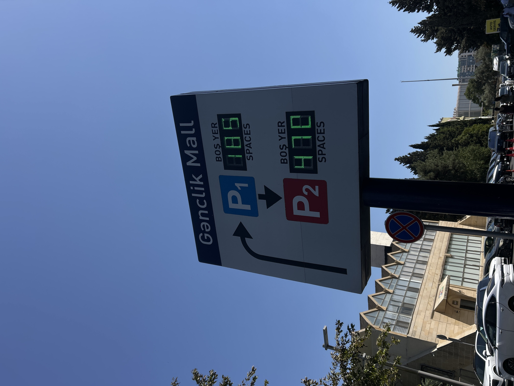 The image shows a clear blue sky above a modern building and a parking lot. A digital sign prominently displays "Gənclik Mall" and provides real-time parking availability for two parking areas, P1 and P2. P1 shows "BOS YER 185 SPACES", indicating 185 available spots, while P2 indicates "BOS YER 470 SPACES", showing 470 available spots. The sign also features directional arrows to guide drivers. In the background, the Gənclik Mall building exhibits a distinctive architectural style with a zig-zag roofline and large glass windows. Various cars are parked in the lot, and a few people are visible walking. A traffic sign with a red circle and a blue cross is mounted on a pole. Further in the background, trees and other buildings suggest an urban environment. The presence of "Gənclik Mall" on the sign and the architectural style of the building strongly suggest the location is Gənclik Mall in Baku, Azerbaijan.