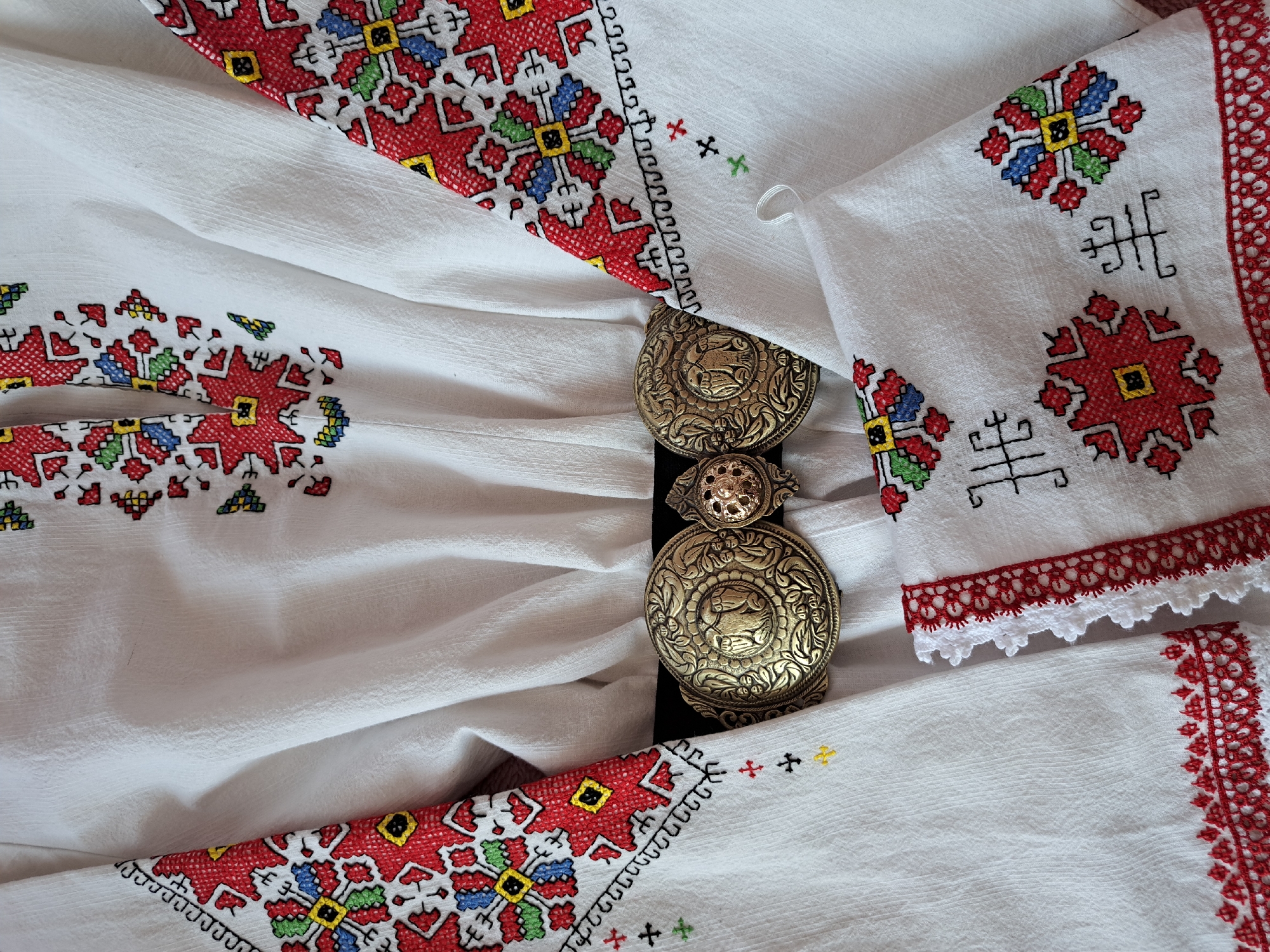 The image displays a close-up of traditional white fabric, intricately embroidered and accented with a decorative metal belt buckle. The white textile, possibly linen or cotton, features a subtle texture and soft folds, suggesting it is part of a garment or an antique textile piece.
The fabric is richly adorned with colorful cross-stitch embroidery. Predominant colors include vibrant red, blue, green, yellow, and black. The patterns are largely geometric and symmetrical, featuring star-like motifs, squares, and abstract shapes. Red is a significant color, forming large patterns and bordering sections. One section of the fabric is finished with a delicate red and white crocheted or embroidered lace trim. Smaller individual cross patterns in black, yellow, and green are also scattered across the white background.
Central to the image is an ornate, antique-style metal belt buckle, appearing to be made of tarnished brass or bronze. The buckle consists of two large, circular components and a smaller connecting piece in the middle, all set against a narrow black band. Each large circular piece is elaborately decorated with raised relief, showcasing a central animal motif surrounded by intricate floral and scrollwork designs. The upper large piece features what appears to be a bird or winged creature, while the lower large piece shows a quadruped, possibly a lion or similar animal. The smaller central piece also bears a detailed floral or starburst pattern.
The overall scene highlights the fine craftsmanship and cultural artistry embedded in traditional textile and metalwork. No people are visible, and no specific activity is taking place. There is no visible text.