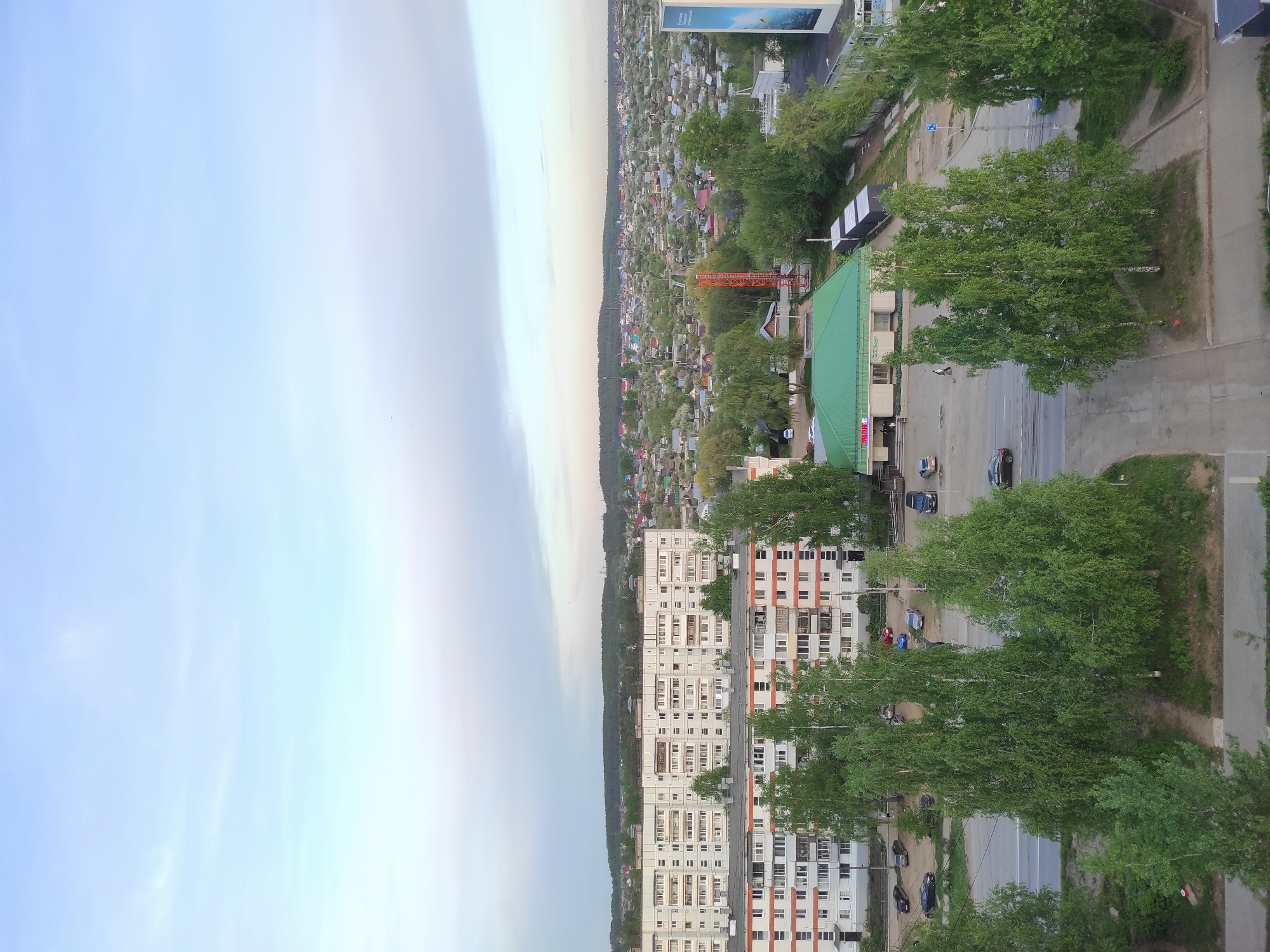 An aerial view of a residential area in Chaikovsky, Russia. The image is oriented vertically. The left side of the image depicts a sky with light clouds. Moving from left to right, there's a treeline and then dense residential housing and garden plots.
In the lower right of the image, a multi-story residential building stands alongside a road lined with trees and sidewalks. Cars are visible on the road and parked alongside the building. A commercial building with a green roof can be seen near the residential area. An advertising billboard is visible near the top of the image.