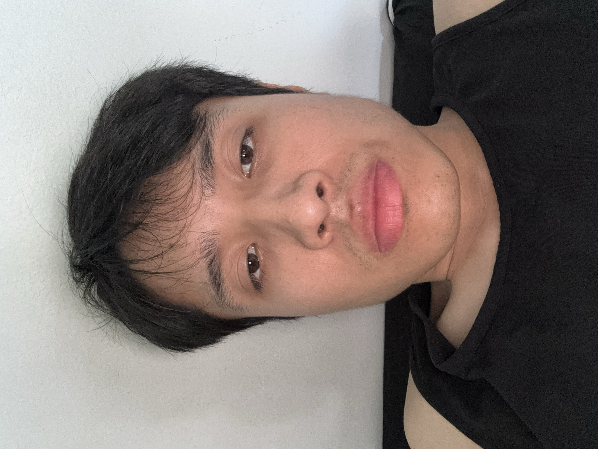 A head-and-shoulders selfie of a young man with dark hair is vertically oriented. He is wearing a black tank top. The background is a plain, light-colored wall. There is no indication of location or city.