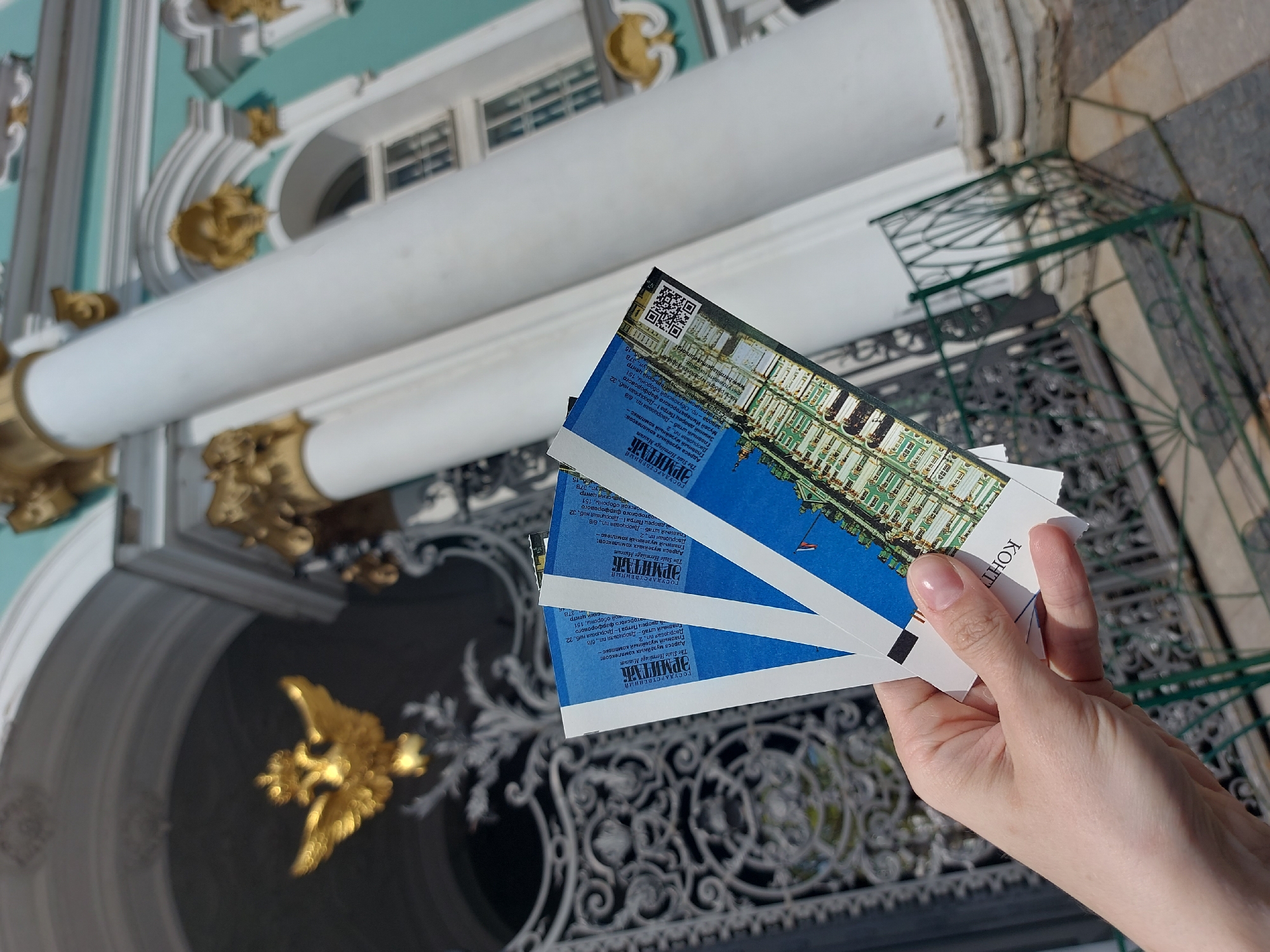 A hand holds several tickets in front of the Winter Palace in Saint Petersburg, Russia. The tickets feature a blue and white color scheme and the name "ЭРМИТАЖ" (Hermitage) in Russian. The Winter Palace, with its ornate architectural details, including columns, gold accents, and decorative ironwork, forms the backdrop. The weather appears bright and sunny.