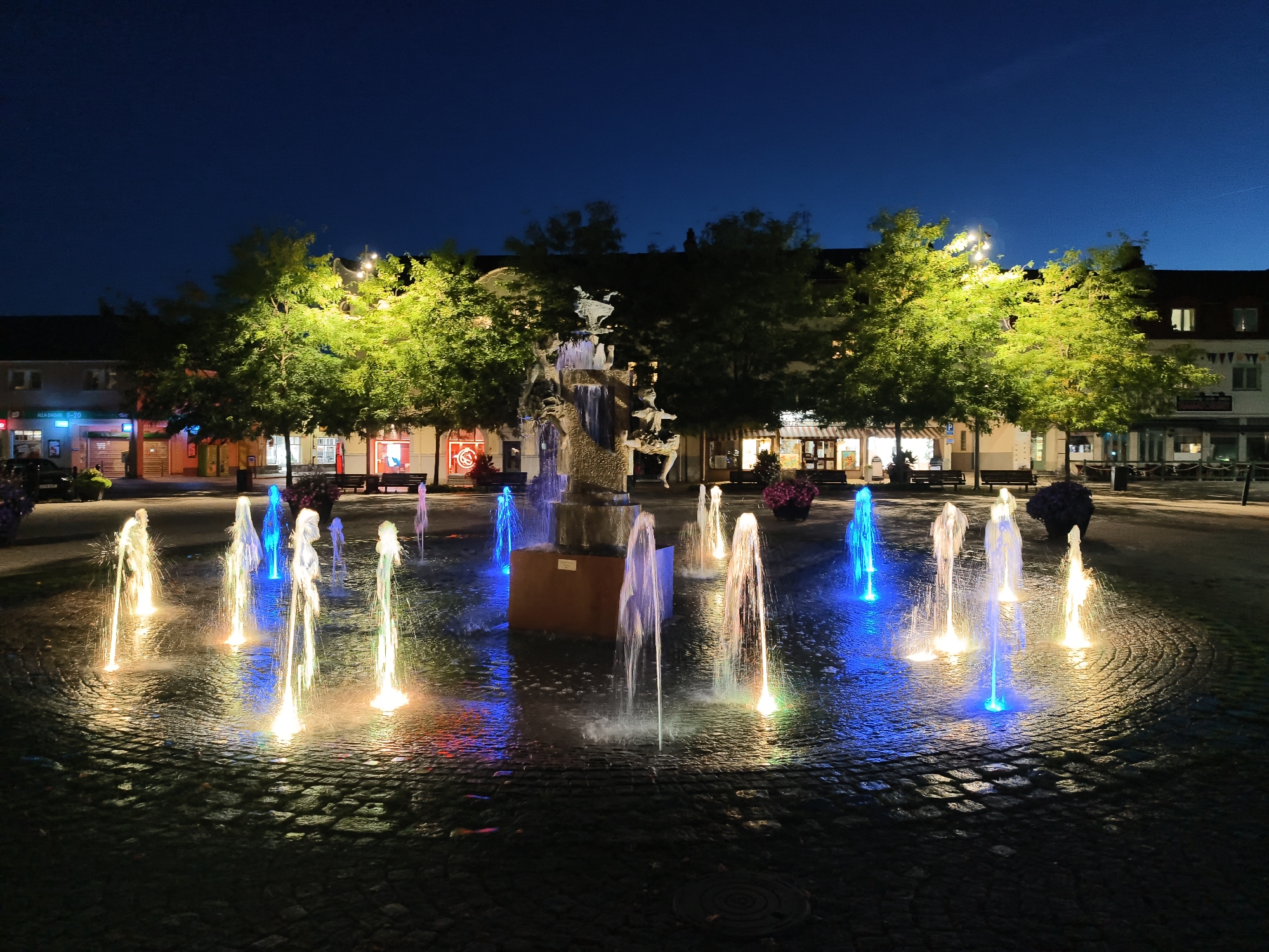 This is a night-time, wide-angle, eye-level, outdoor photograph of a lit public fountain with multiple water jets in Söderköping, Sweden. The fountain is located in a paved public square, which is lined with trees that are illuminated by artificial lights. In the background, buildings with shops and windows are visible. The fountain itself is the primary subject, with illuminated water jets of varying heights and colors, including white, blue, and yellow. The water reflects on the dark, cobblestone-like pavement. A central sculpture is visible behind the water jets, though it is partially obscured. There are no people visible in the immediate vicinity of the fountain, although some benches and possibly trash receptacles are present in the background. The sky is a deep, dark blue, indicating it is nighttime.