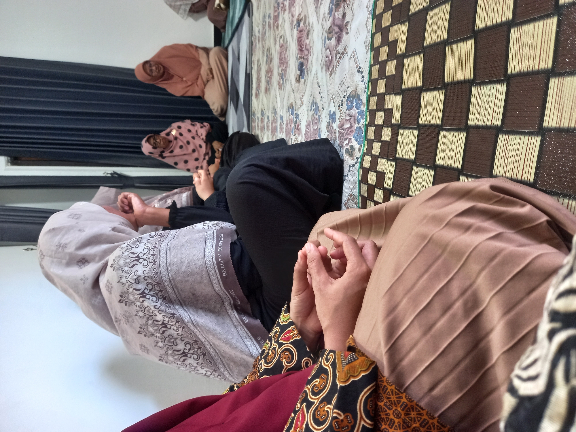 The image shows a group of women sitting on the floor, possibly in a home or community space in Tempos, Indonesia. The women are wearing modest clothing, including headscarves (hijabs). The woman in the foreground on the left is wearing a maroon top and a patterned skirt, with her hands clasped in her lap. To her right, another woman is wearing a light gray hijab with a patterned border. In the background, several other women are visible, some wearing patterned headscarves and others in solid colors. The floor is covered with a patterned rug and a woven mat with a checkerboard design. Dark blue curtains are visible in the background. The lighting suggests an indoor setting, possibly during the daytime.