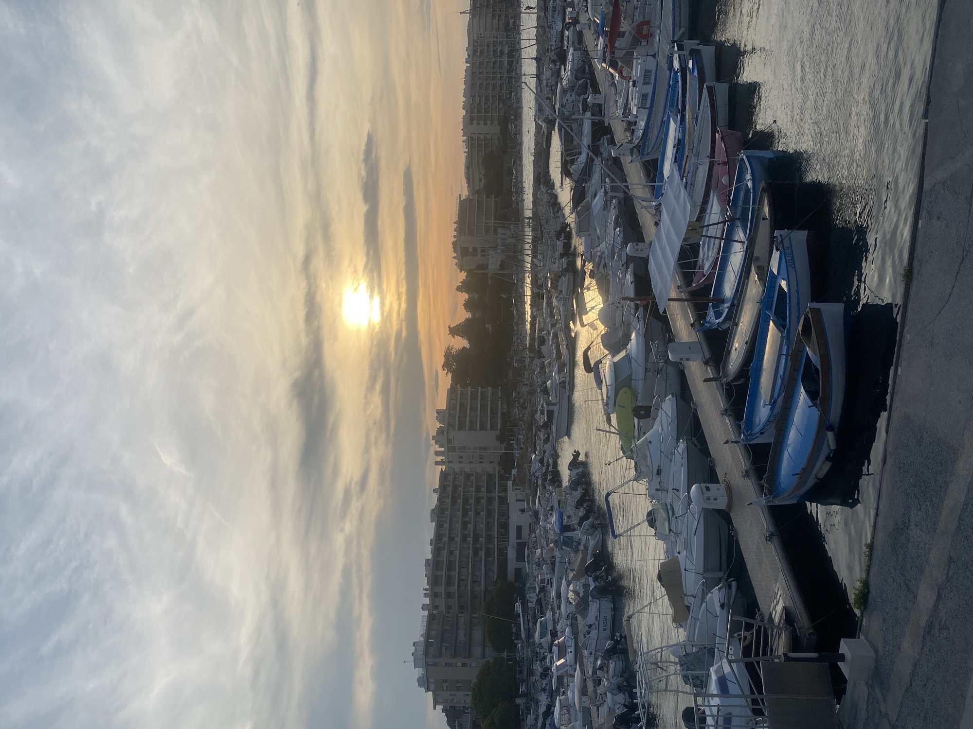 This image captures a harbor in Antibes, France, likely during sunset. The sky is filled with soft, wispy clouds, with the sun peeking through, casting a warm, golden glow over the scene. The harbor is densely packed with a variety of boats, ranging from small dinghies to larger motorboats. Many of the boats are moored along wooden docks, and their reflections shimmer on the water's surface. In the background, several multi-story buildings, characteristic of coastal towns, line the waterfront. The overall atmosphere is tranquil and serene, suggesting a peaceful evening in a Mediterranean port.