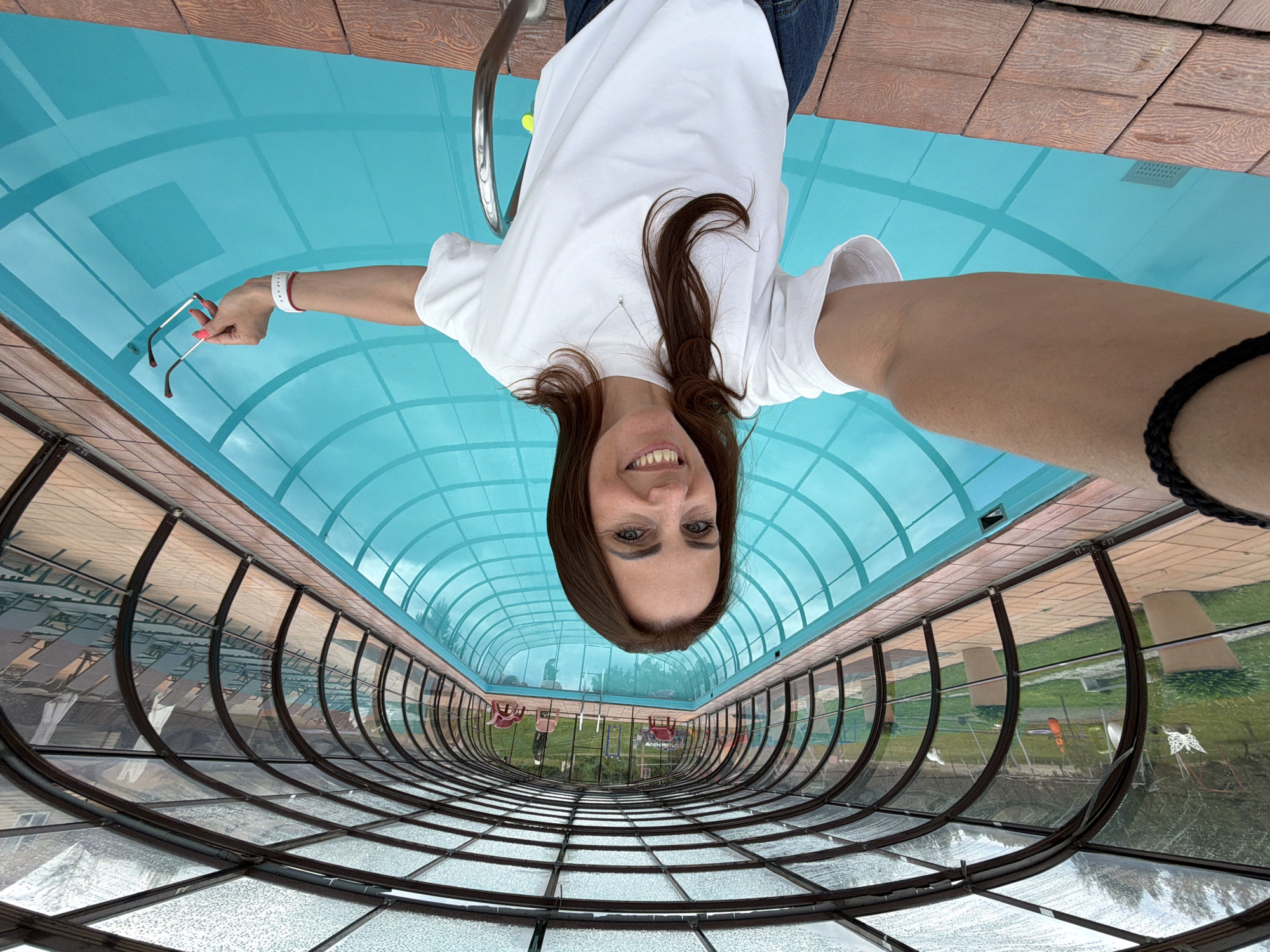 A woman with brown hair is taking a selfie in an indoor swimming pool. She is wearing a white t-shirt and denim shorts. She is holding sunglasses in her right hand. The pool is covered by a transparent roof with a metal frame. The pool area is surrounded by wooden decking. In the background, there is a green lawn with trees and some lawn chairs.