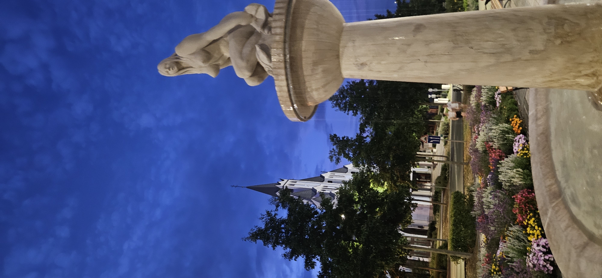 A vertical, low-angle shot captures a serene evening scene in Hévíz, Hungary. In the foreground, a concrete fountain features a nude female statue crouching on its rim. The statue is made of stone and has long hair. The fountain is situated in front of a vibrant flowerbed filled with purple, pink, yellow, and white blossoms, with a manicured green hedge behind them.
Beyond the flowerbed, a row of trees lines a street, with a white church featuring a tall, dark steeple visible in the background. The sky above is a deep blue with scattered clouds, indicating twilight. Two figures, appearing to be young adults, walk on the sidewalk to the right of the flowerbed, their backs to the camera. The scene is illuminated by ambient light, creating a peaceful and tranquil atmosphere.