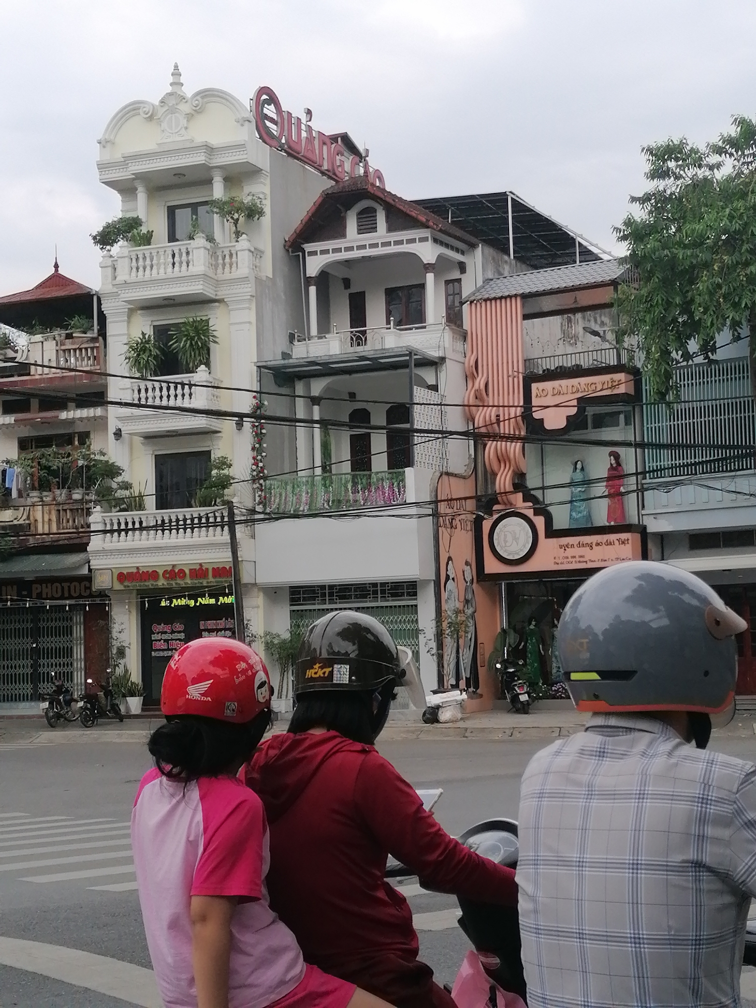 The image depicts a street scene in Lao Cai Ward, Vietnam. Three people on a motorbike are in the foreground. The person driving wears a red hoodie and a dark helmet. The person behind the driver wears a pink shirt with a red helmet. The person in front of the driver wears a plaid shirt and a grey helmet.
Behind them, a street lined with buildings and shops is visible. On the left, there is a building with a sign that says "IN-PHOTOC" and "QUẢNG CÁO HÀI". Another building features a sign that reads "QUANG CAO." Next to it, there is a building with mannequins dressed in traditional Vietnamese attire, "ÁO DÀI DÀNG VIỆ". Some motorbikes are parked along the side of the street. The sky is overcast. Overhead power lines run across the street.