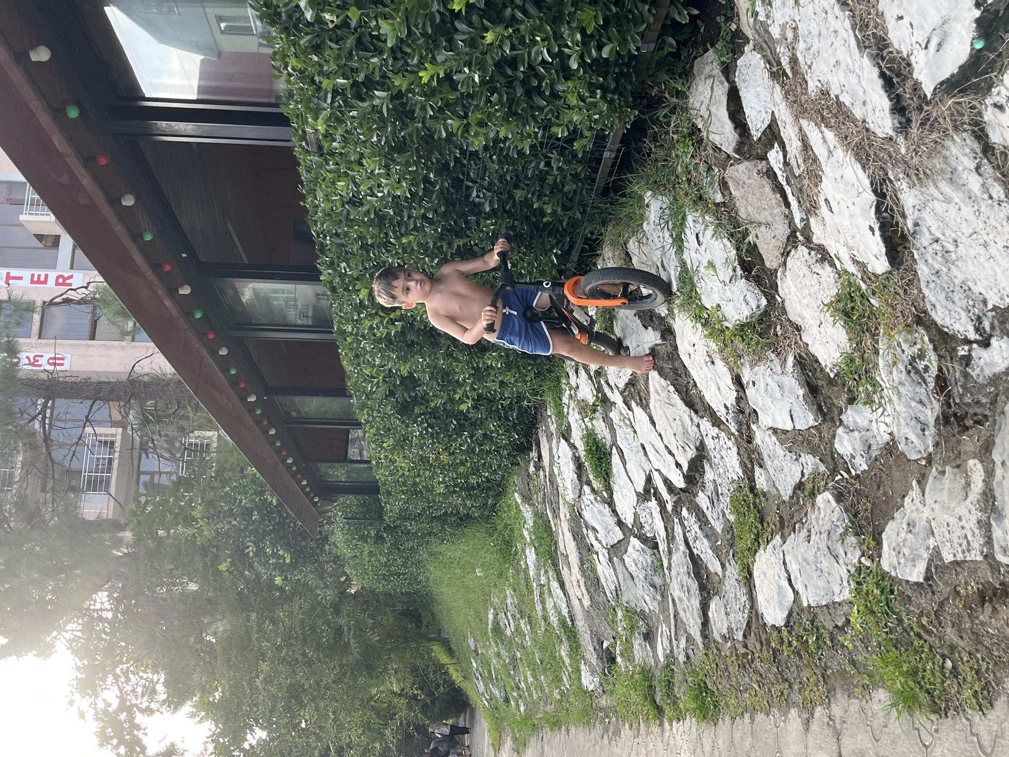 A young boy, shirtless and wearing blue shorts, rides a small orange and black balance bike on a pathway made of flat, irregularly shaped gray stones. He is barefoot and looks to his left. The pathway is on a slope, with green grass growing between the stones. To the right of the pathway is a dense green hedge, and behind the hedge is a building with large windows, topped by a dark brown overhang decorated with red and green lights. A few trees are visible behind the building, and the sky is bright with sunlight filtering through the branches. Further left, a paved sidewalk is visible, with a few people walking in the distance. The scene appears to be outdoors in a recreational area, possibly near a cafe or restaurant, given the building and its decorations. This location is identified as being in the city of Ureki, Georgia.