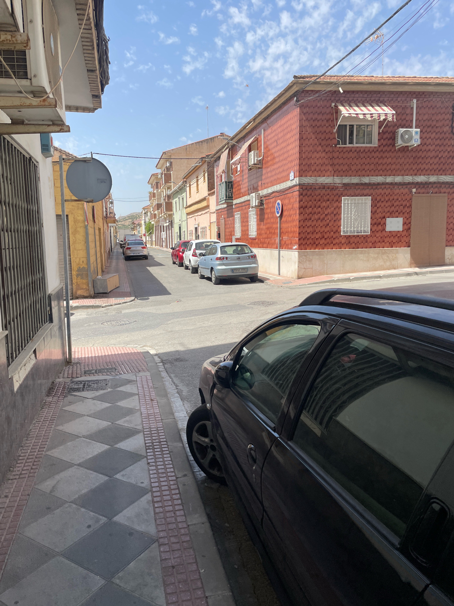 This is a street-level, eye-level, medium shot of a street scene in Atarfe, Spain. The street is lined with buildings on both sides. The building on the right is a two-story structure with a red tiled facade and awnings over the windows. The building on the left is lighter in color and has a metal grate covering a window. Several cars are parked along the street. A black car is prominently featured in the foreground, on the right side of the image. The sidewalk in the foreground has a checkered tile pattern. The sky is blue with scattered clouds. The scene appears to be during daylight hours.