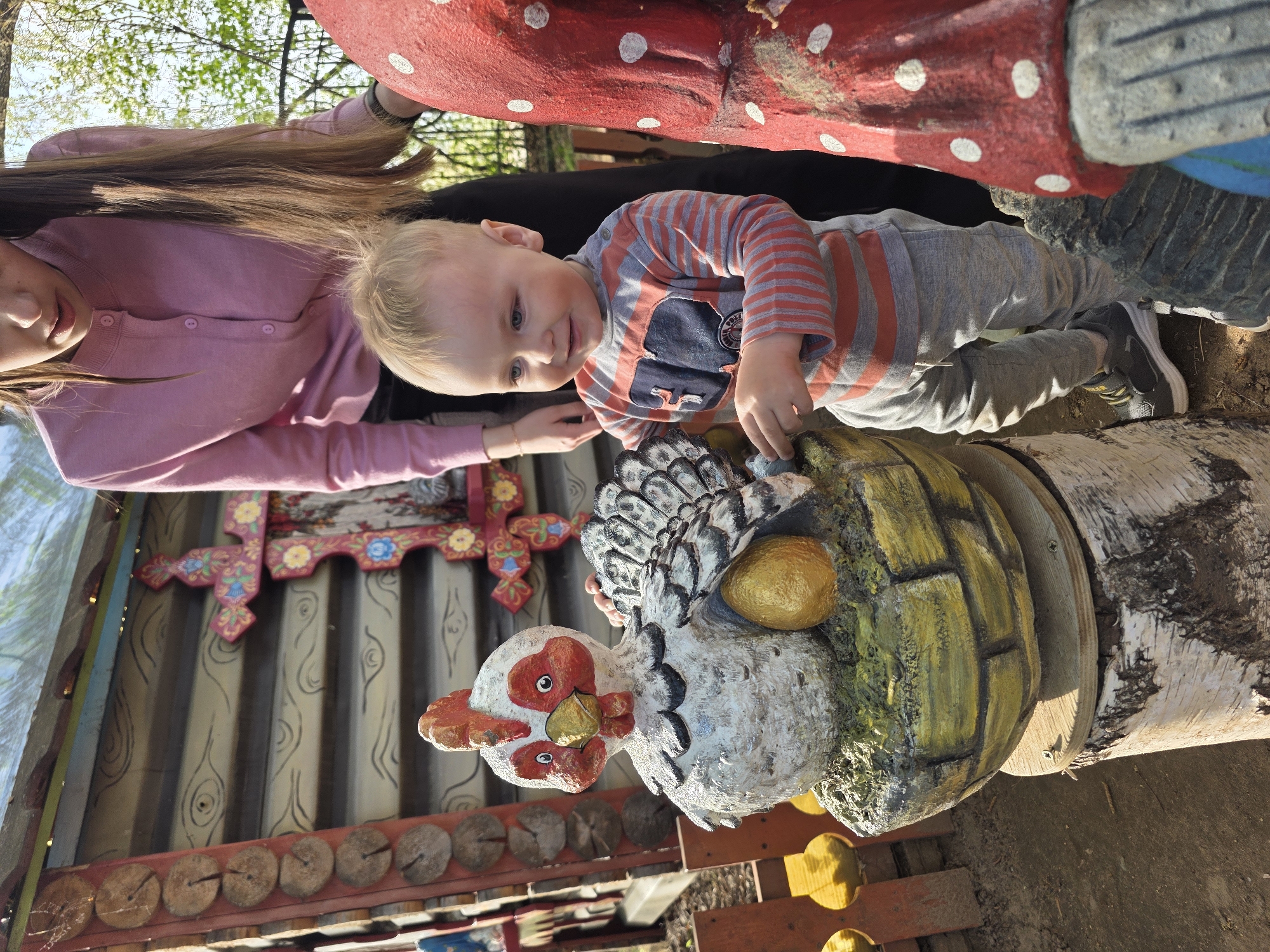 The image shows a young child in a striped shirt and gray pants standing next to a painted concrete sculpture of a hen. A woman's hand rests near the child. The hen sculpture is detailed with a red and white head, a golden beak, and white and gray feathers. It also has a single golden egg. The sculpture sits atop a cylindrical base made to look like a well constructed of yellow bricks. This well base is attached to a birch log.
Behind the child and sculpture is a part of a building or structure with a decorative cross affixed to it. The background also contains green foliage. The location is Chaikovsky, Russia.