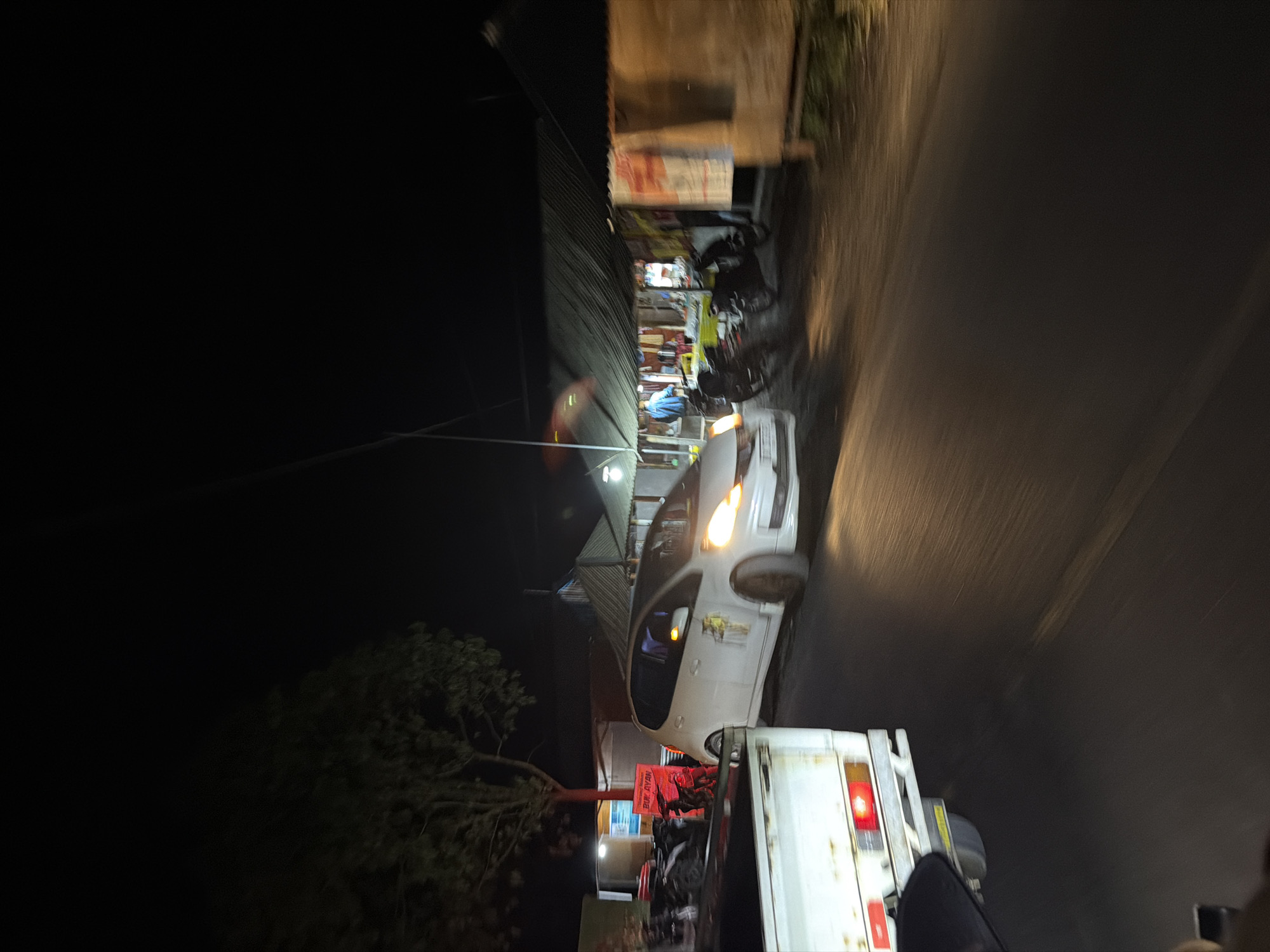 This is a low-light, motion-blurred photograph taken at night, likely in Kintamani, Indonesia. The scene depicts a roadside setting with several vehicles and what appears to be a small market or food stalls.
In the foreground, the back of a white truck is visible, with its red taillight illuminated. To the left of the truck, a white car is parked or moving slowly. The car has its headlights on and appears to be a compact model. Behind the car and to the left, a covered market stall or building with a corrugated metal roof is visible. Various items, possibly merchandise or food, are displayed. Some people are present near the stalls, with one individual wearing a blue shirt.
Several motorcycles are parked or in motion near the stalls, suggesting a busy area. A large tree with dark foliage is on the far left of the image. The road surface is visible, appearing wet or reflecting light from the vehicles and stalls. The overall impression is of a vibrant nighttime scene on a roadside.
There is text visible on a red banner in front of the stalls, partially obscured, which reads "BUKA JAYAN". The time of day is clearly night, and there are no obvious cues about the weather, though the road surface suggests it might have rained recently or is damp. The motion blur indicates movement, either from the vehicles or the photographer.