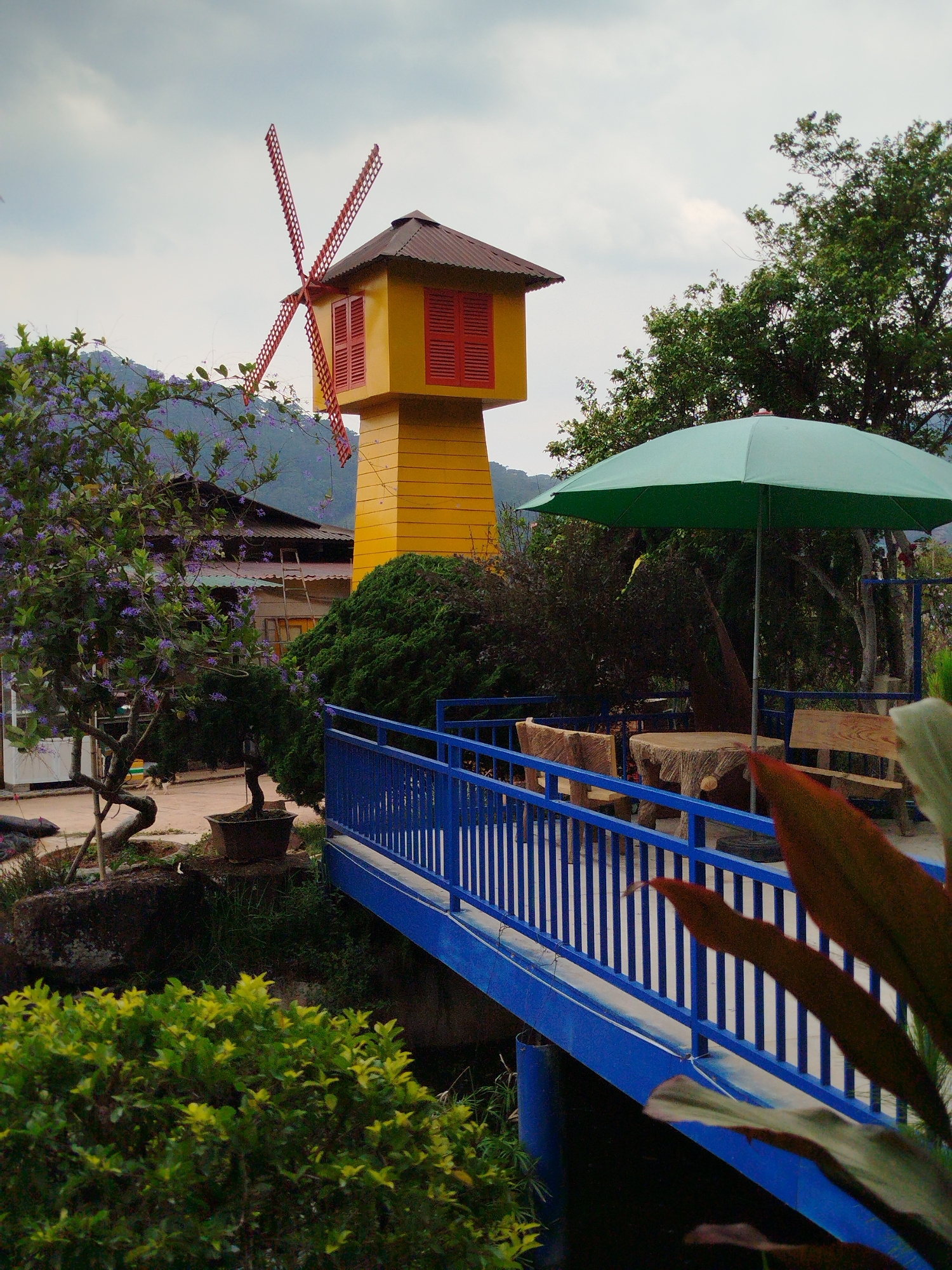 Captured in Da Lat, Vietnam, the image presents a landscape marked by distinct man-made structures. A yellow tower, capped with a red windmill and shuttered windows, rises prominently on the left side of the frame. Adjacent to it, a blue bridge spans a small waterway. A green umbrella stands nearby, shading a rustic-looking picnic table set. Lush greenery frames the scene, with a tree bearing small purple flowers in the foreground. The sky is overcast.