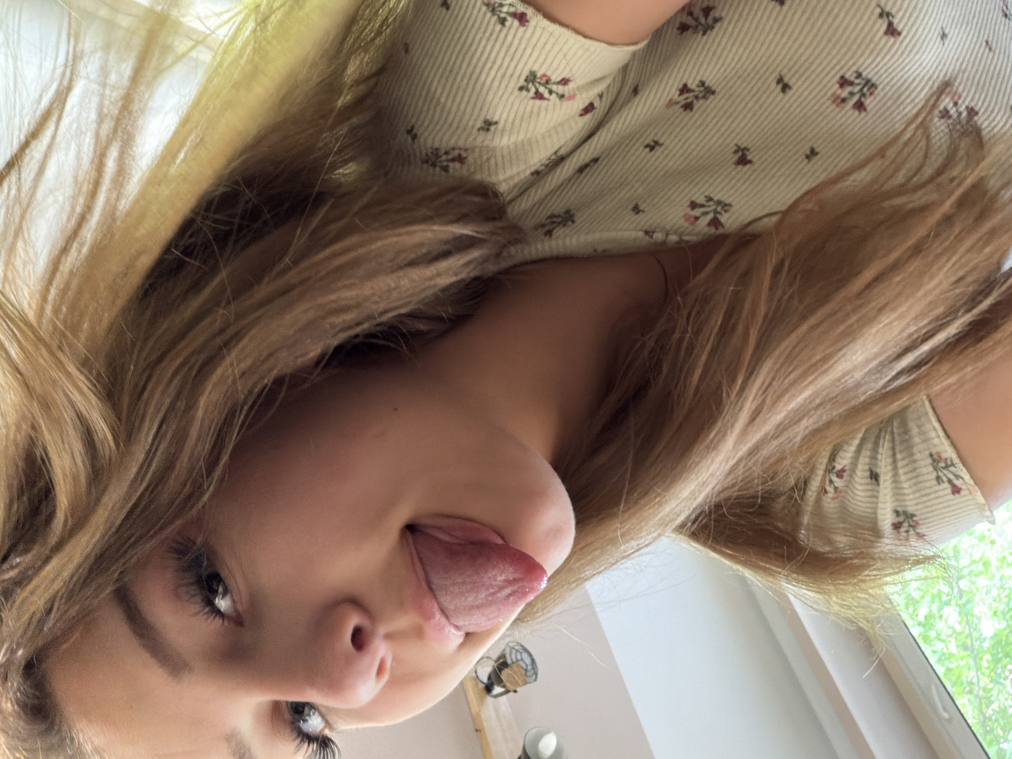 This close-up, low-angle selfie features a young woman with light brown to blonde hair, playfully sticking her tongue out. Her head is tilted, giving the impression of looking up from below, with her face partially framed by her loose hair. Her eyes, adorned with visible eyelashes, are directed towards the camera, conveying a casual and mischievous expression.
She is wearing a light-colored, possibly off-white or cream, ribbed top that features a subtle, repetitive pattern of small floral designs in shades of red, pink, and green. Her long hair cascades around her face and shoulders, adding to the informal feel of the image.
The background reveals an indoor setting with a bright, light-toned ceiling or upper wall. To the right, a window offers a glimpse of lush green foliage outside, suggesting trees or bushes and indicating natural daylight. Another indistinct object, possibly a light fixture or small fan, is visible further in the background. The lighting suggests it is daytime. The overall mood is one of lightheartedness and playfulness. No visible text is present in the image. This scene is set in Banya, Bulgaria.