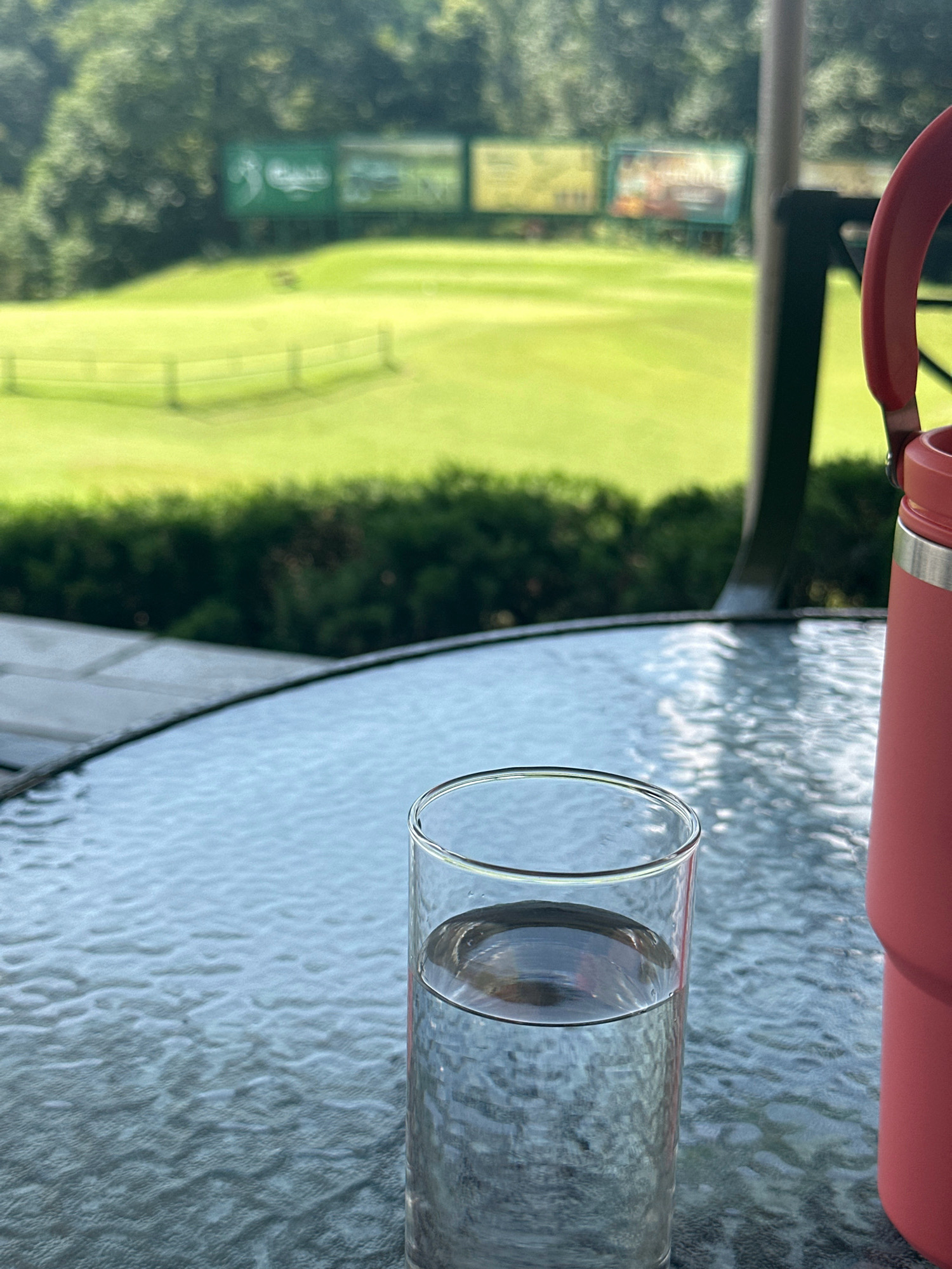A close-up, eye-level shot depicts a glass of clear water and a coral-colored insulated tumbler on a textured glass table. The tumbler is positioned to the right, with its handle visible. In the background, a lush green golf course stretches out, featuring manicured lawns, a distant fence, and several blurred billboards. Trees form a dense backdrop, and a dark railing separates the table area from the golf course. The lighting suggests an outdoor, daytime setting. The image is not located in Kageshwori Manohara, Nepal.