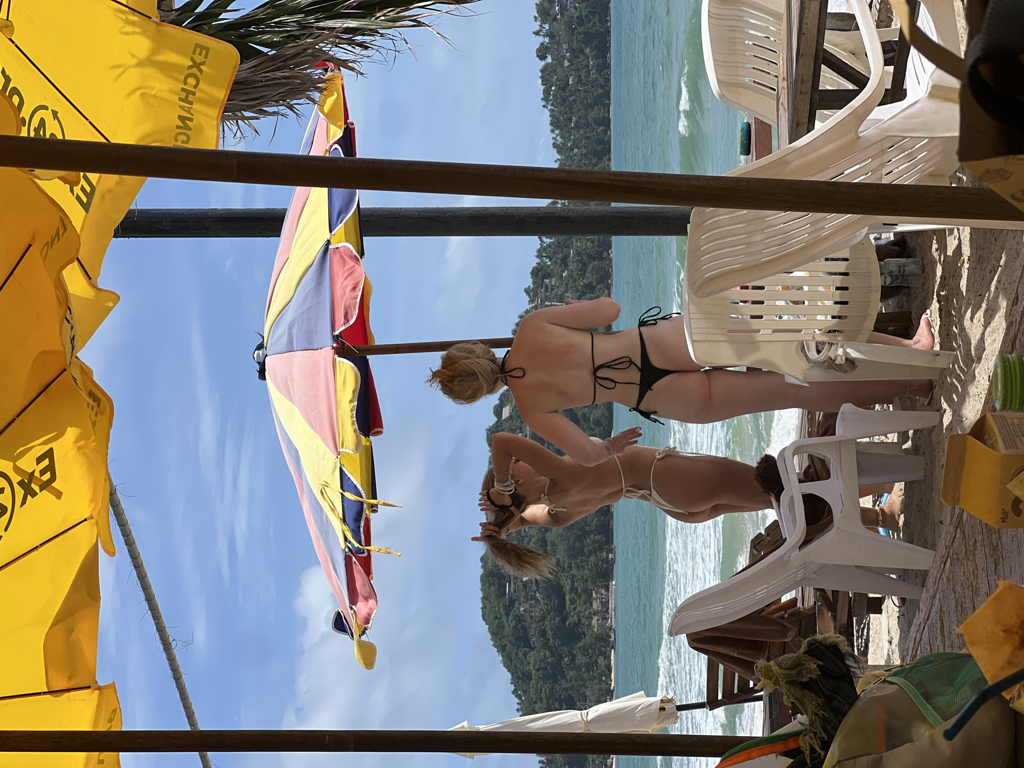 Two women in bikinis stand on a sandy beach under a colorful beach umbrella. The woman on the left, facing away from the camera, has blonde hair pulled into a ponytail and is wearing a light-colored string bikini. She is adjusting her hair. The woman on the right, also facing away, has blonde hair tied up and is wearing a black string bikini. They are standing in front of the ocean, with waves visible. In the background, a green, tree-covered hill rises from the water, with some buildings visible on its slopes. There are white plastic chairs and wooden tables in the foreground. The sky is blue with scattered clouds, and palm fronds are visible to the right.