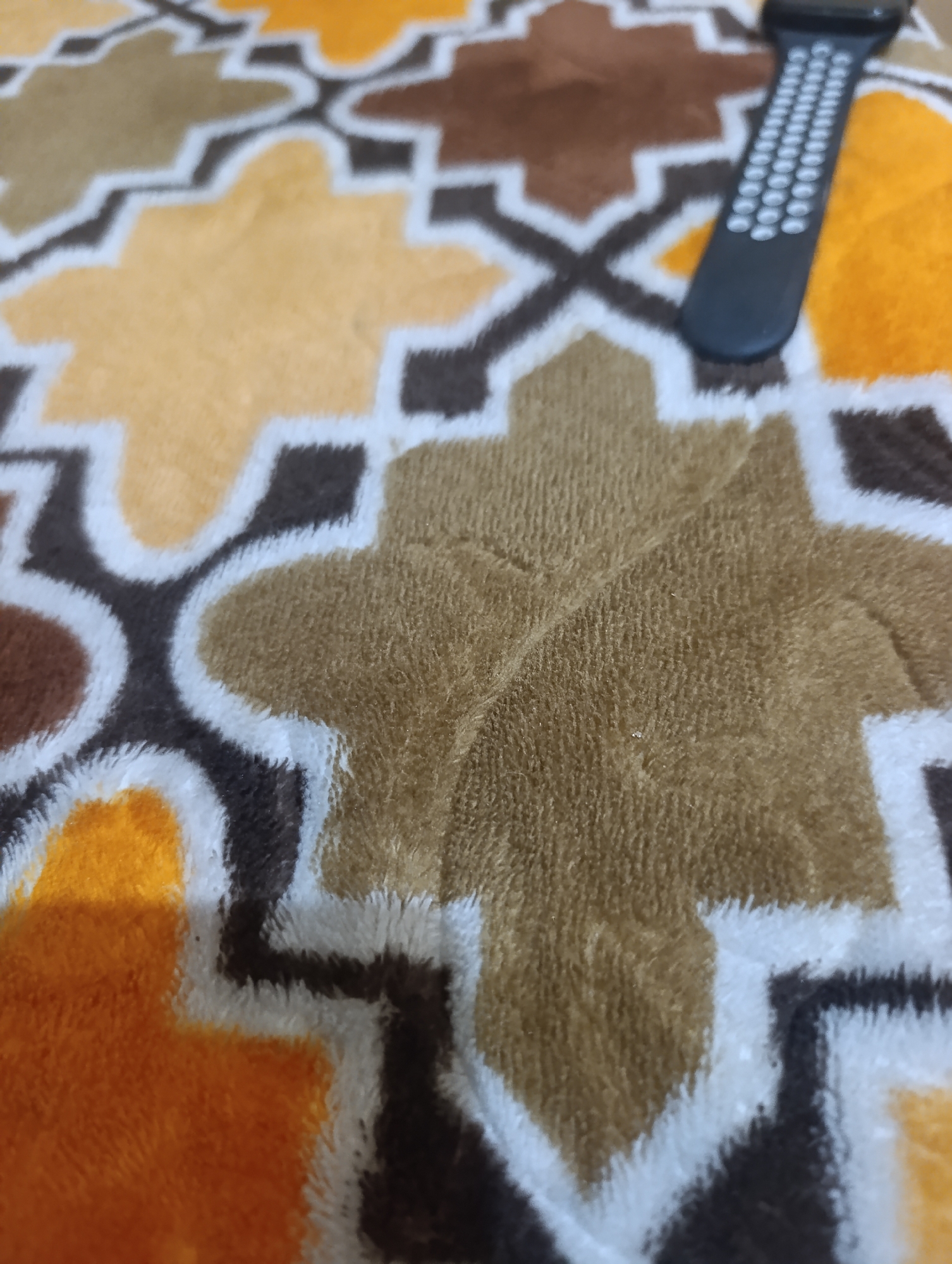 A black smartwatch with a textured band rests on a fuzzy blanket. The blanket features a repeating geometric pattern in shades of orange, brown, and white. The image is a close-up shot, focusing on the texture of the blanket and the details of the smartwatch band. The setting is likely indoors, given the presence of the blanket and smartwatch. No specific location in Madatte, Indonesia is discernible from the image.