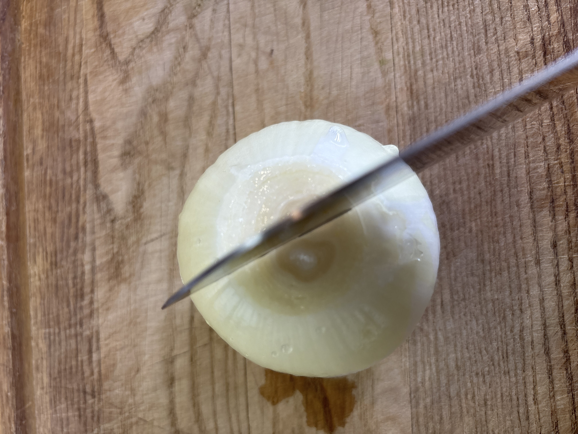 A close-up, overhead shot shows a metal knife cutting into a peeled, white onion on a wooden cutting board. The knife is angled, with its blade partially submerged into the top of the onion. The cutting board has a visible wood grain and appears slightly damp, with a small puddle of liquid to the left of the onion. The background is out of focus. The image provides no information regarding its location in Bila Tserkva, Ukraine.