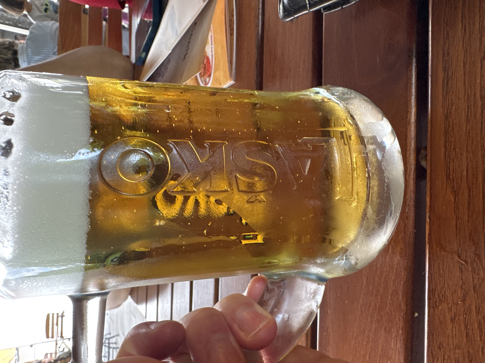The image shows a close-up of a half-full glass mug of golden beer with a thick white foam head, condensation on the outside of the glass, and bubbles rising within the liquid. The beer mug has a raised logo or text that appears to say "LJUBSKO". A person's hand is visible on the left side of the mug, holding its handle. The mug rests on a wooden table. In the background, out of focus, are several people sitting at tables, some chairs, and a menu or papers on the table. The setting appears to be an outdoor seating area of a restaurant or cafe. Based on the logo, it is possible this is from a brewery or establishment in Slovenia. The city of Postojna, Slovenia, is famous for its caves and castle, but this image depicts a common scene at a beer garden or restaurant within the country.
