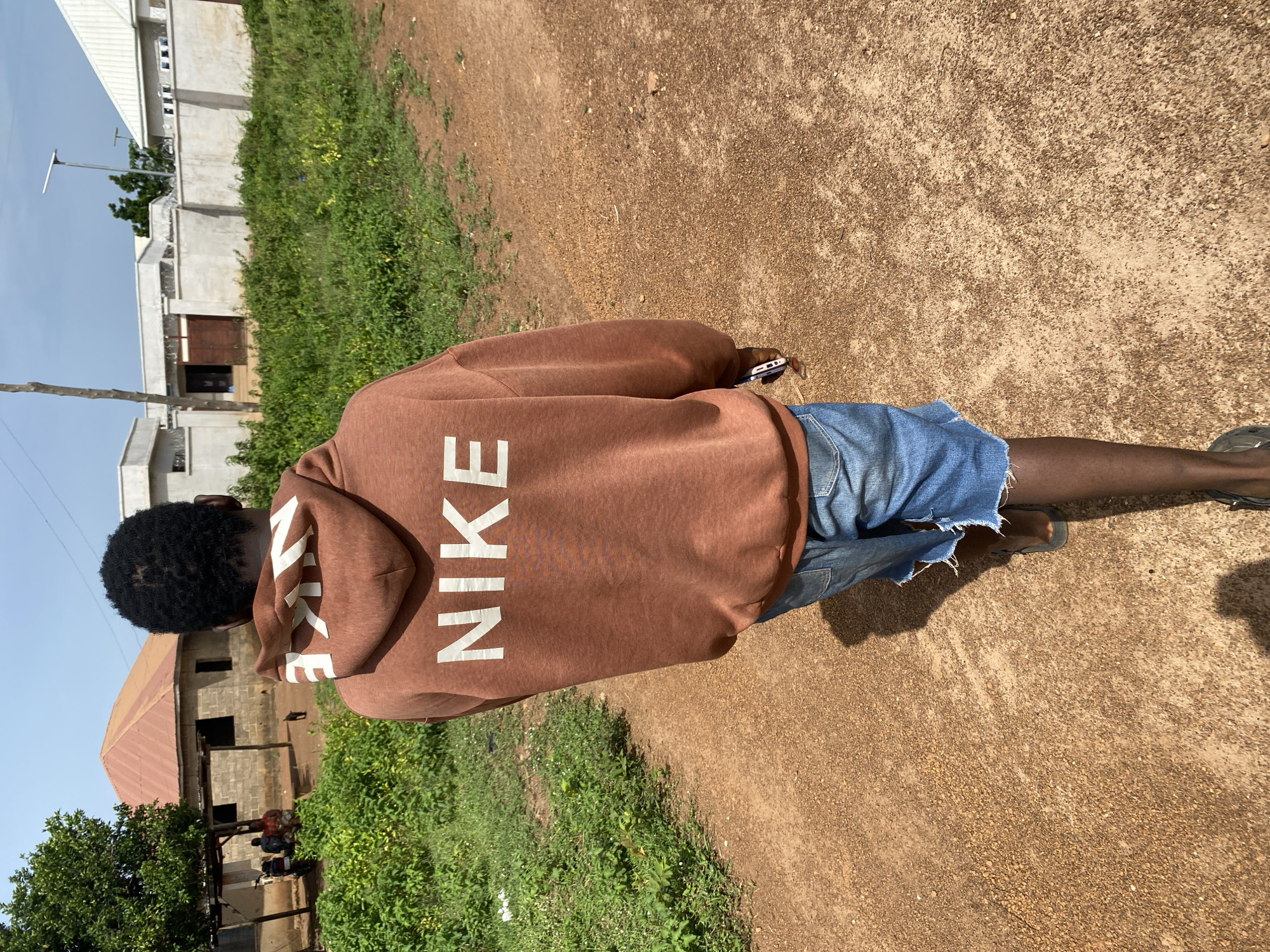 A person walks away from the camera on a dirt path. The person is wearing a brown Nike hoodie and denim shorts. The person is holding a phone in their right hand. The path is surrounded by green grass and some buildings in the background. The sky is blue and cloudy.