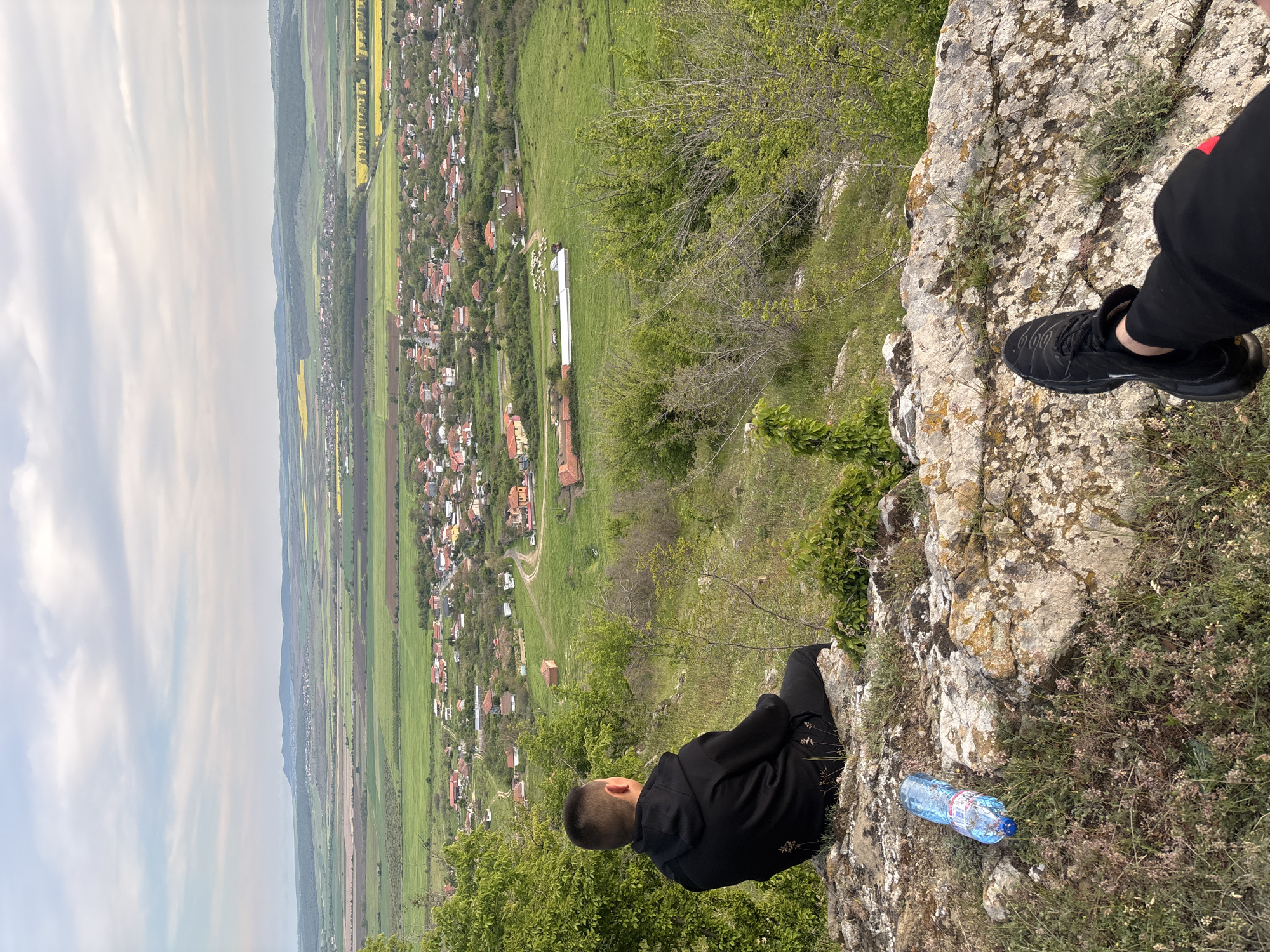 The image depicts a person sitting on a rocky ledge overlooking a valley landscape. The person is wearing a black hoodie and black pants. To the right of the person is a black shoe and a water bottle. The valley contains a village with houses, green fields, and a long building. The sky is overcast. The image is taken from a high vantage point. Although there is no visible sign for the city, the location is Goritsa, Bulgaria.