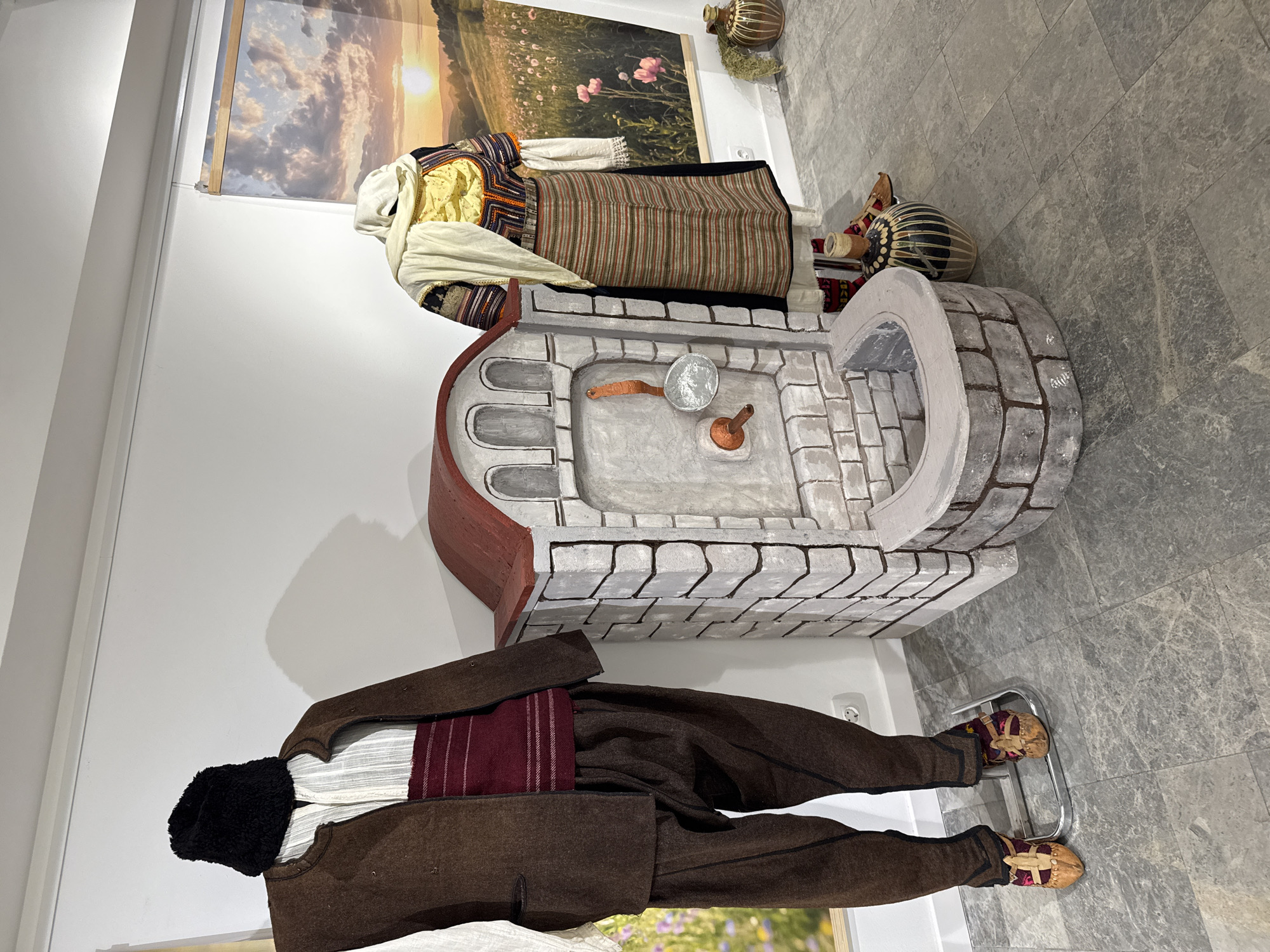 The image depicts a display of traditional Bulgarian clothing and artifacts. On the left, a mannequin is dressed in a brown vest, white shirt, red sash, and brown trousers, with a black fur hat. On the right, another mannequin is adorned in a colorful embroidered blouse, a striped skirt, and a white headscarf. Between the mannequins is a miniature stone fountain with a red roof, featuring a metal ladle. The background includes a large landscape photograph of a sunset over a field with wildflowers. Additional decorative pottery is visible to the right. This display is likely located at the "House of Humor and Satire" in Gabrovo, Bulgaria, known for its exhibitions related to Bulgarian culture and traditions. The overall environment appears to be an indoor exhibition space with light-colored walls and a grey tiled floor.